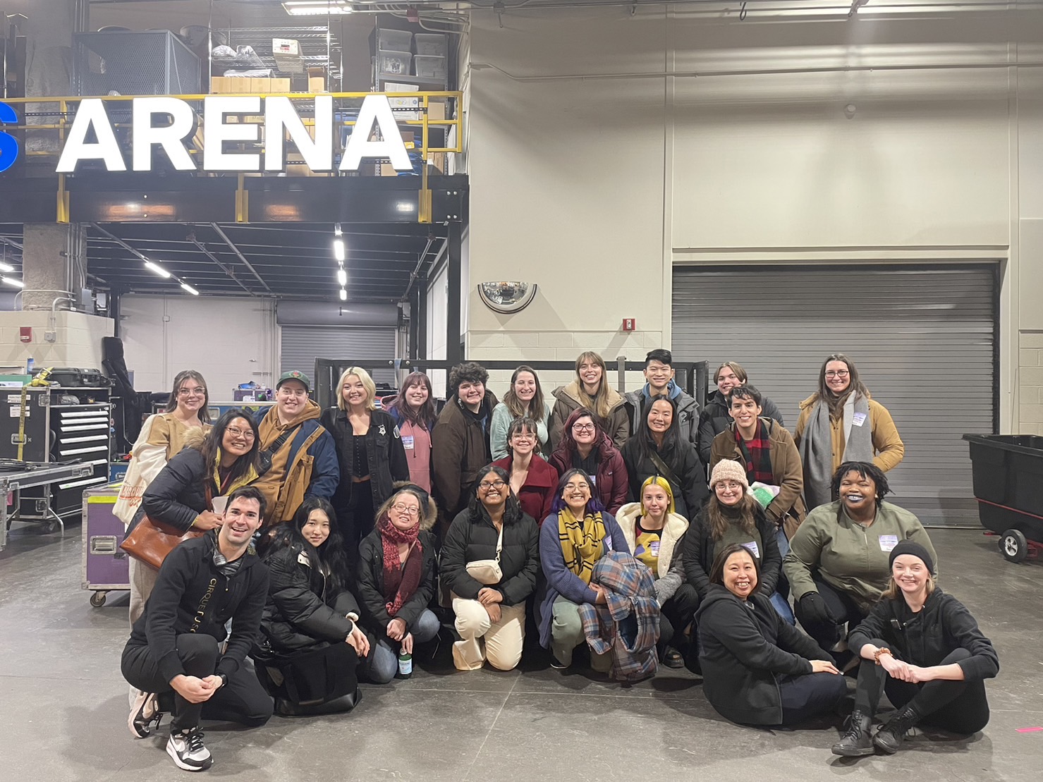 A large group of students pose for a picture, smiling, under a large sign that says ARENA.