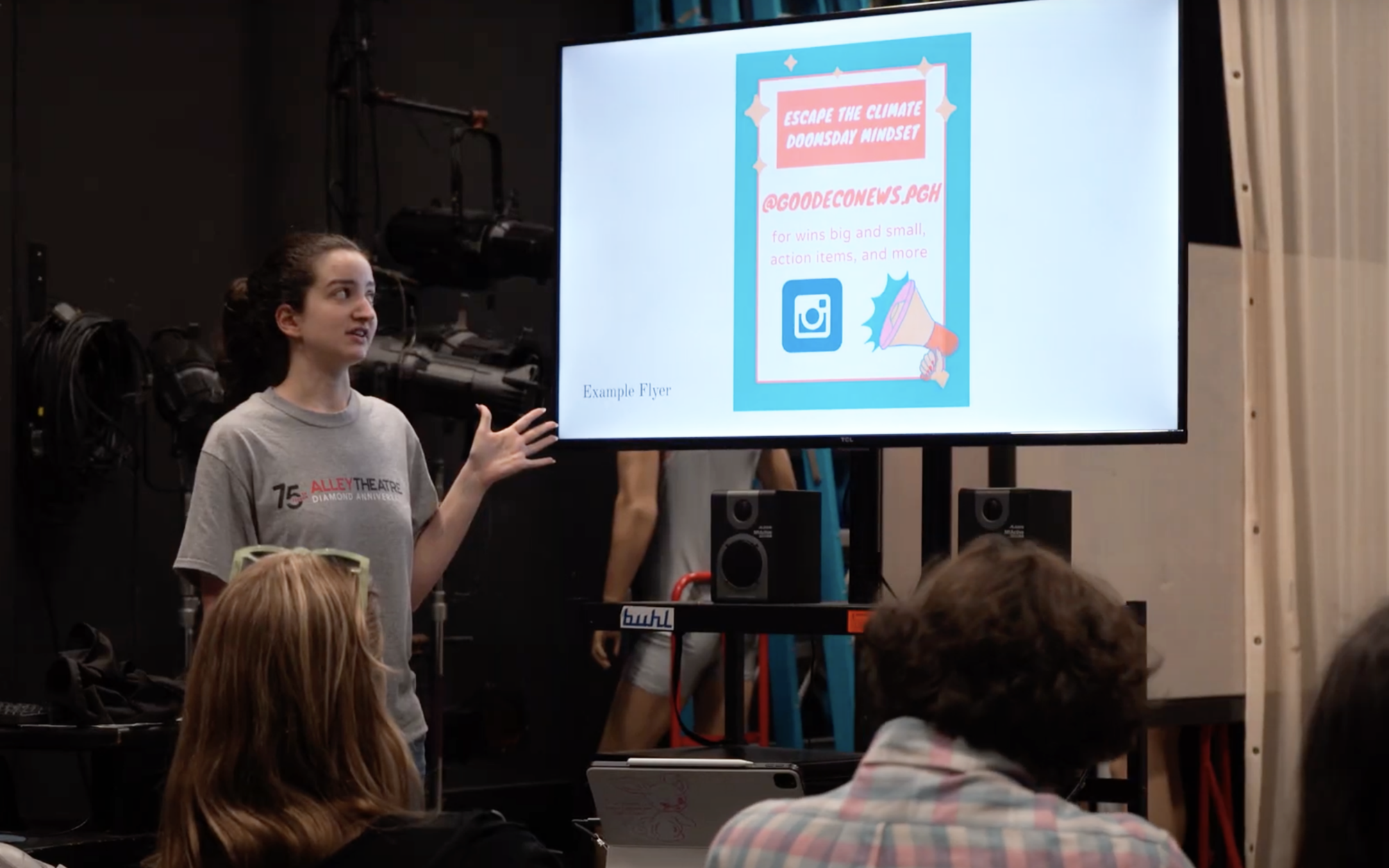 A student wearing a gray t-shirt with her brown hair in a ponytail gestures toward a screen that has a social media post displayed.