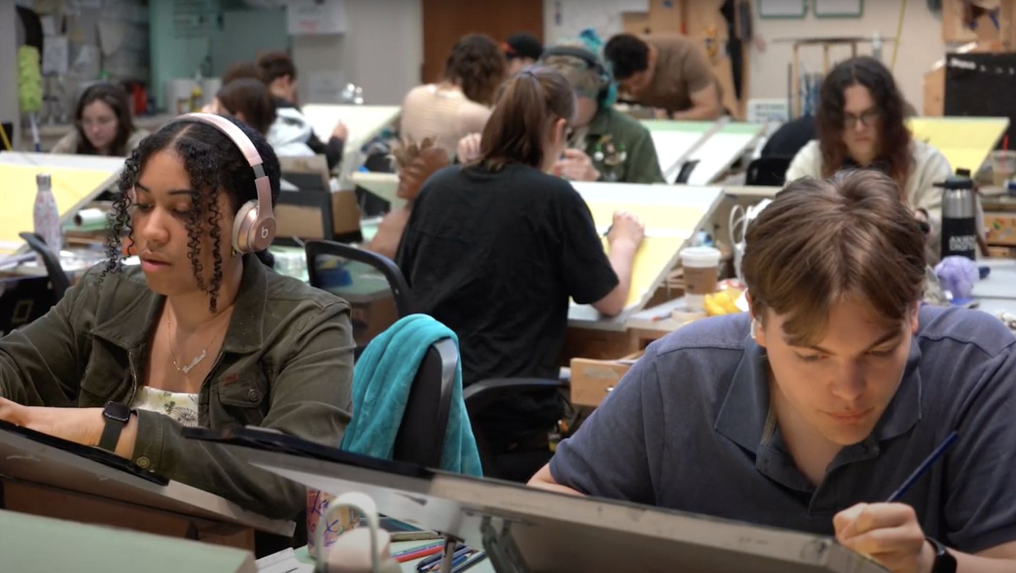 Students sitting at individual drafting stations, facing different directions in a classroom, working on drawing projects.