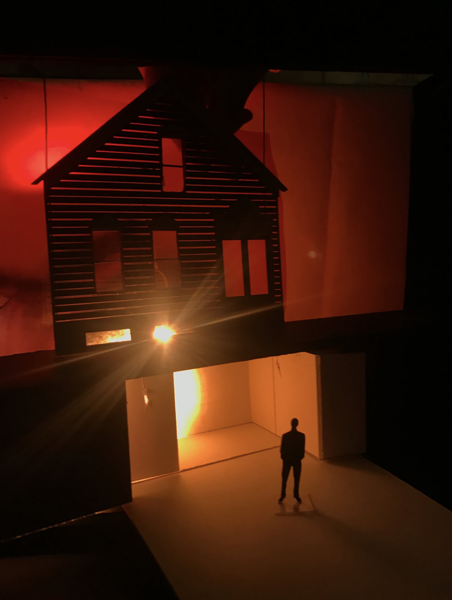 A model set depicting a facade of a house and a bright orange light shining in from behind; a small figure stands in front of the house.