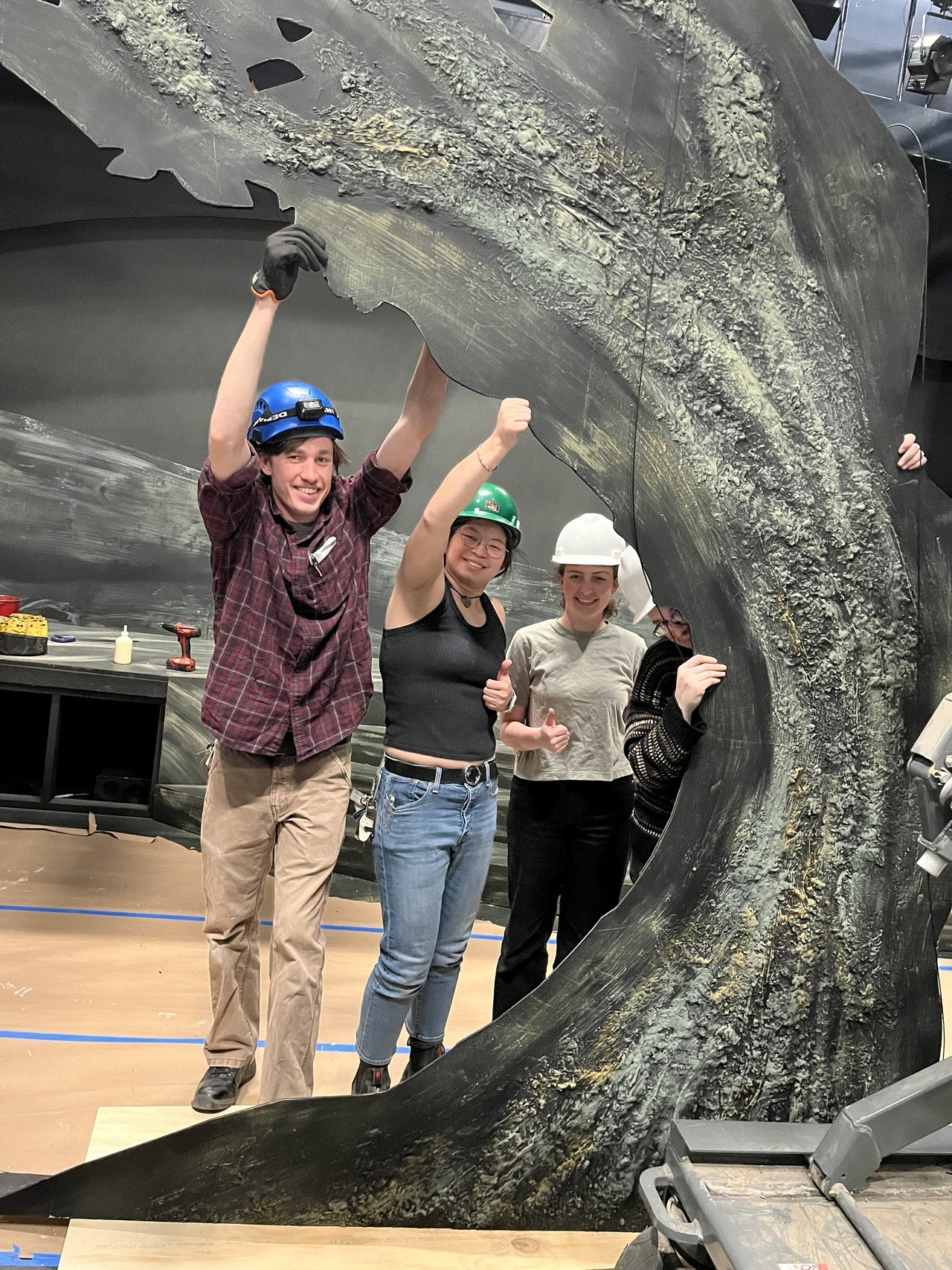 Three students wearing hard hats hold up a large set piece in the Helen Wayne Rauh Studio Theater.