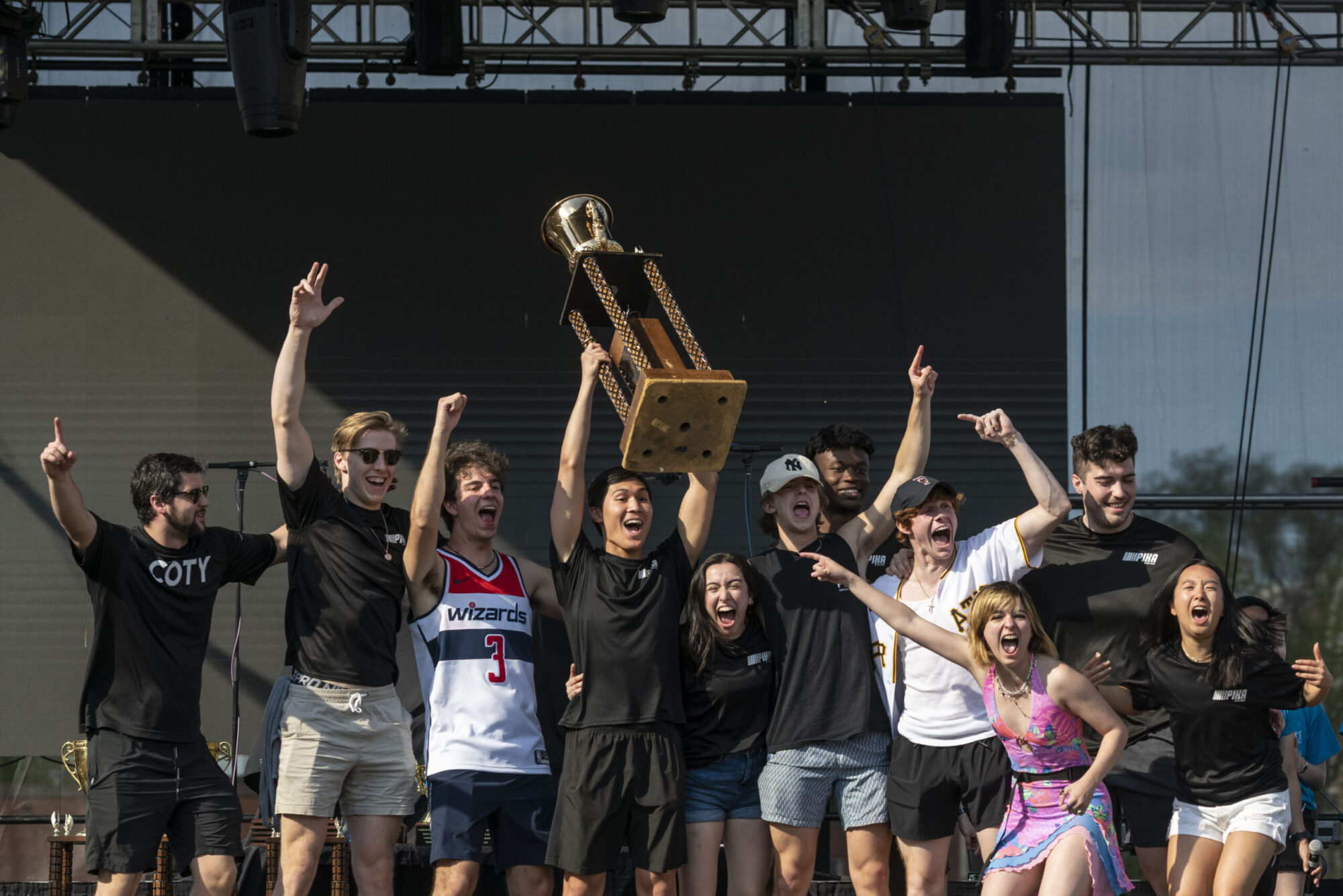 A group of ten students standing on an outdoor stage, cheering and holding up a trophy they won for CMU’s Buggy competition.