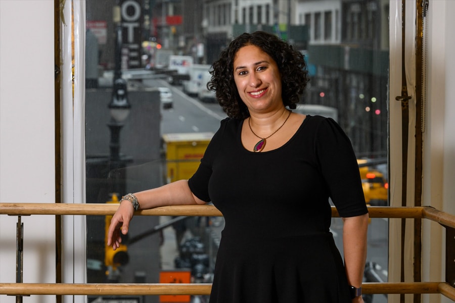 Liz Coleman stands at a dance bar in a studio with a city street out the window behind her; she wears a black dress and smiles at the camera.