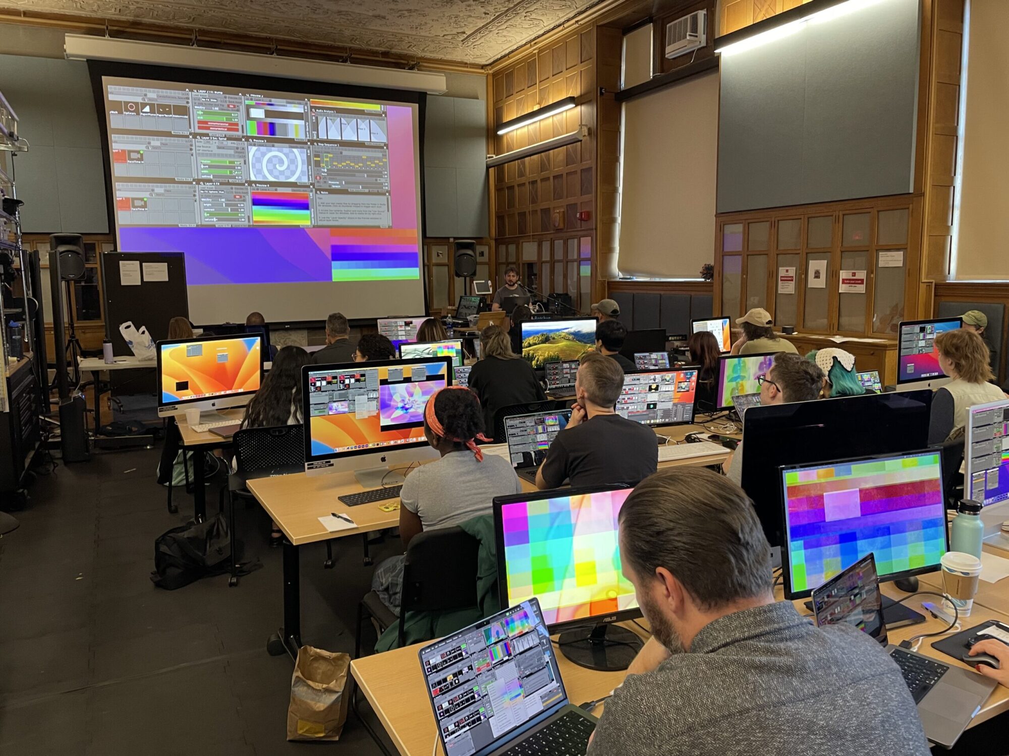 A classroom with a large screen at the front and students at tables working on computers and laptops with screens showing colorful media and technology.