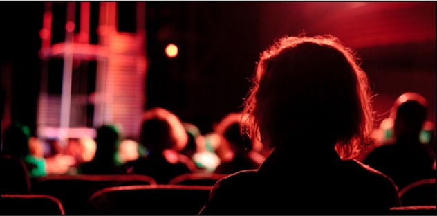 A photo from behind of an audience watching a performance; the space is dark with red lights.