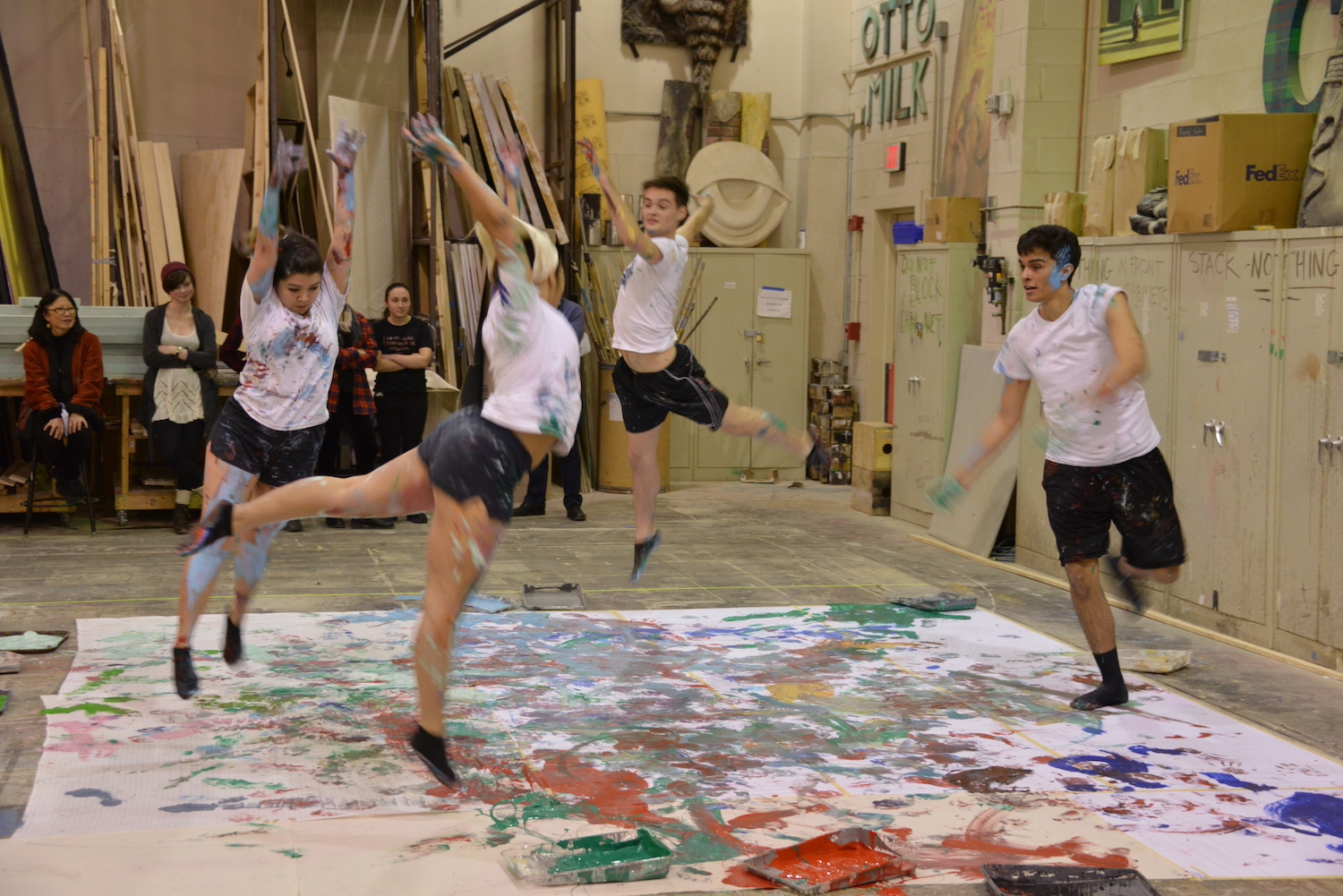 Photo of four students dancing on a paint covered sheet in the School of Drama scene shop.