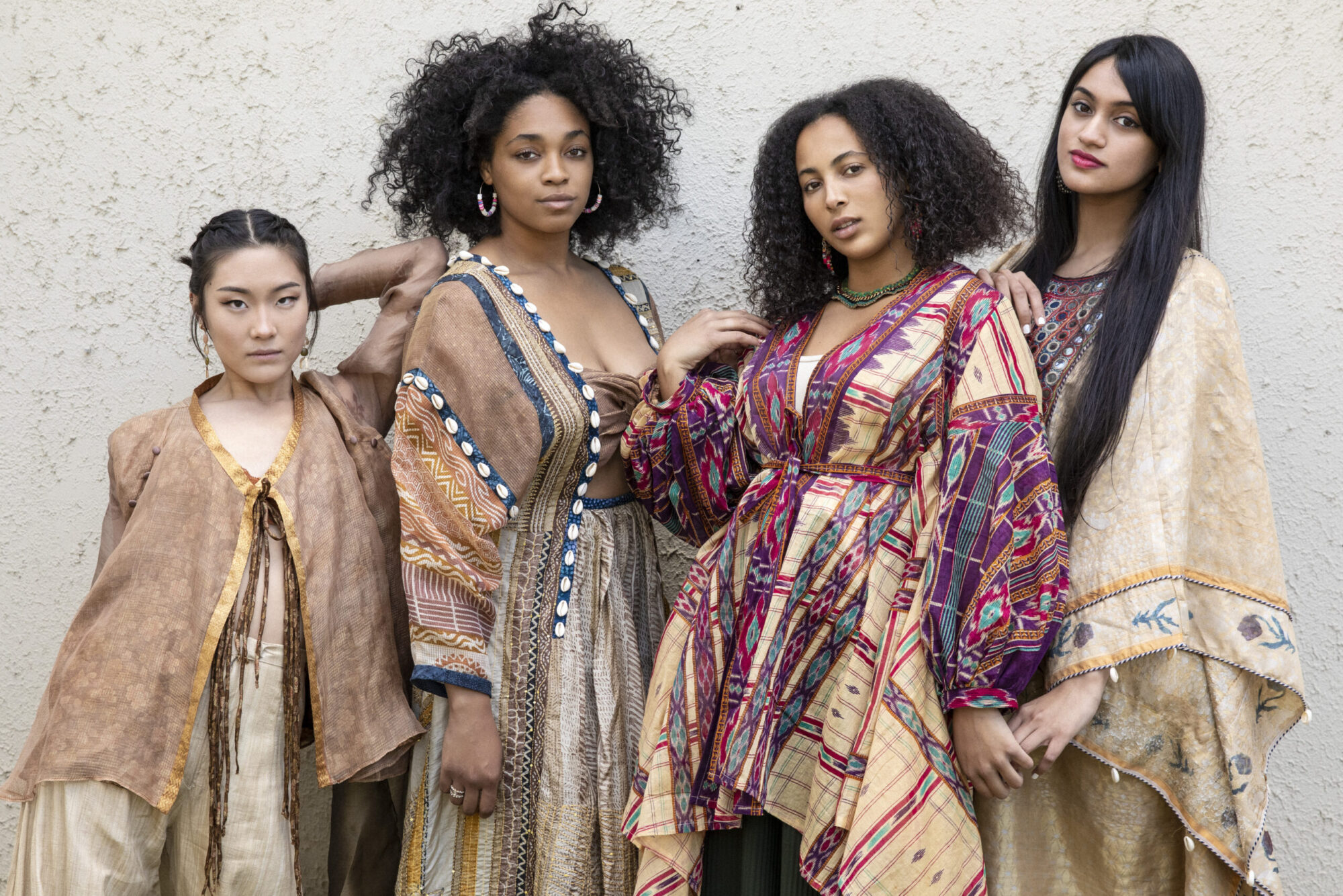 Four women stand wearing intricate costumes made of neutral color fabric with detailed color accents in pinks and blues.