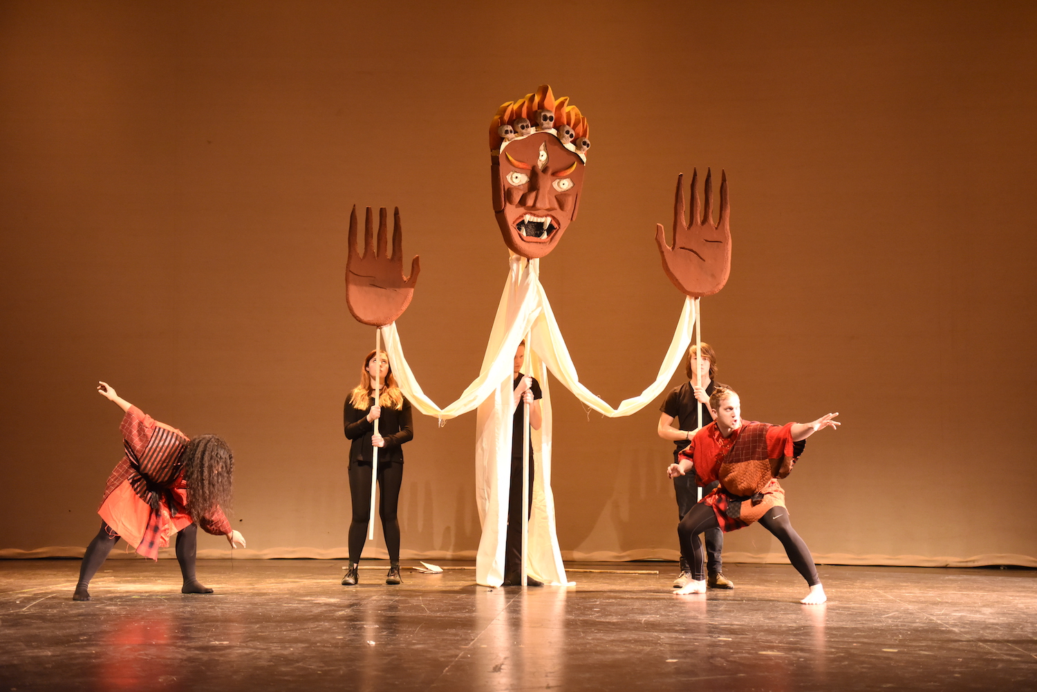 A giant puppet on stage, maneuvered by three people, with a giant terracotta mask and hands; two performers in red frocks dance on either side of it.