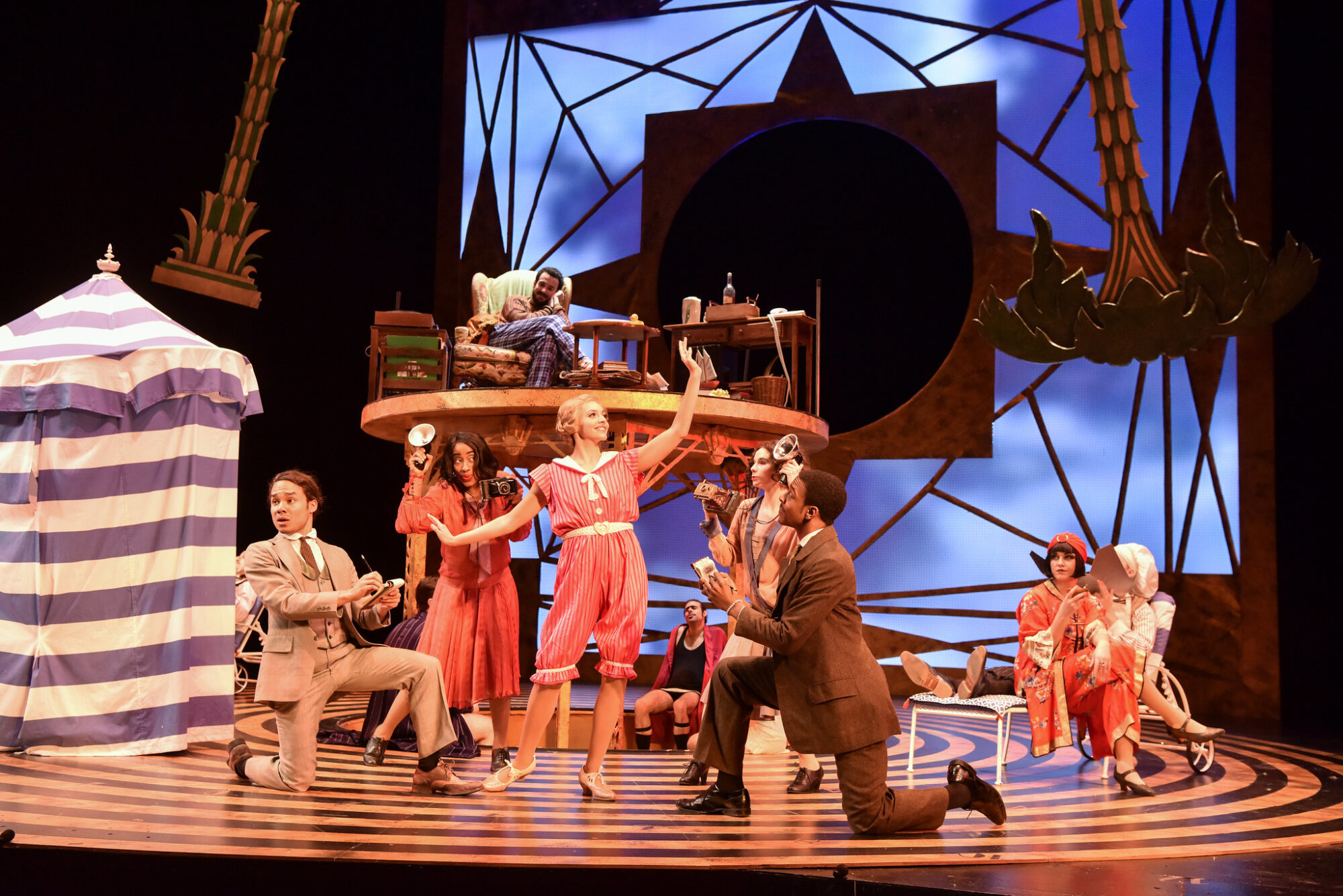 An elaborate stage scene with a concentric circle design on the floor, actors posing on stage, and an elevated living room scene suspended from above, where another actor watches the scene below.