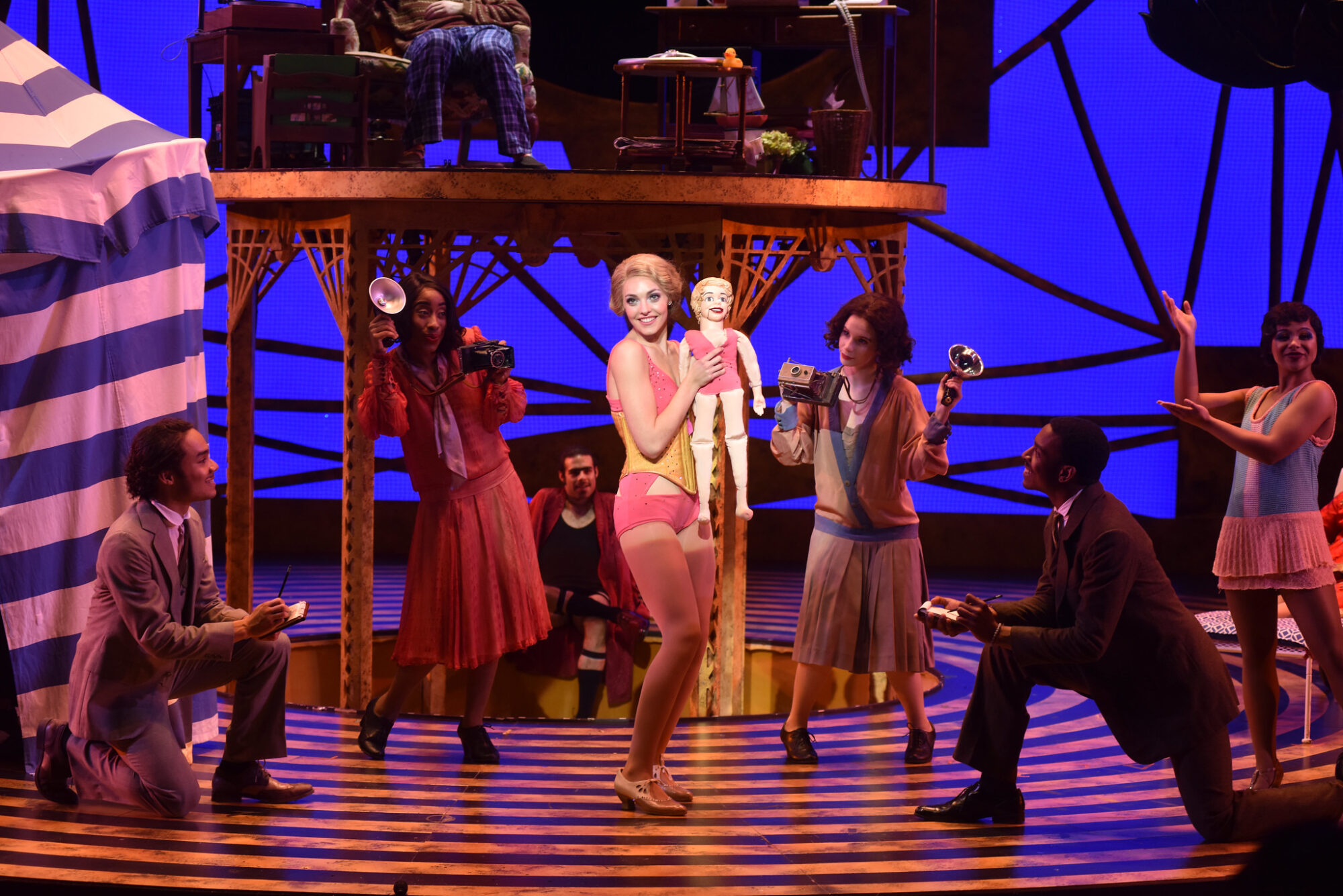 An actor is in a spotlight on stage, holding a ventriloquist doll, surrounded by other actors as reporters in a scene from “The Drowsy Chaperone.”