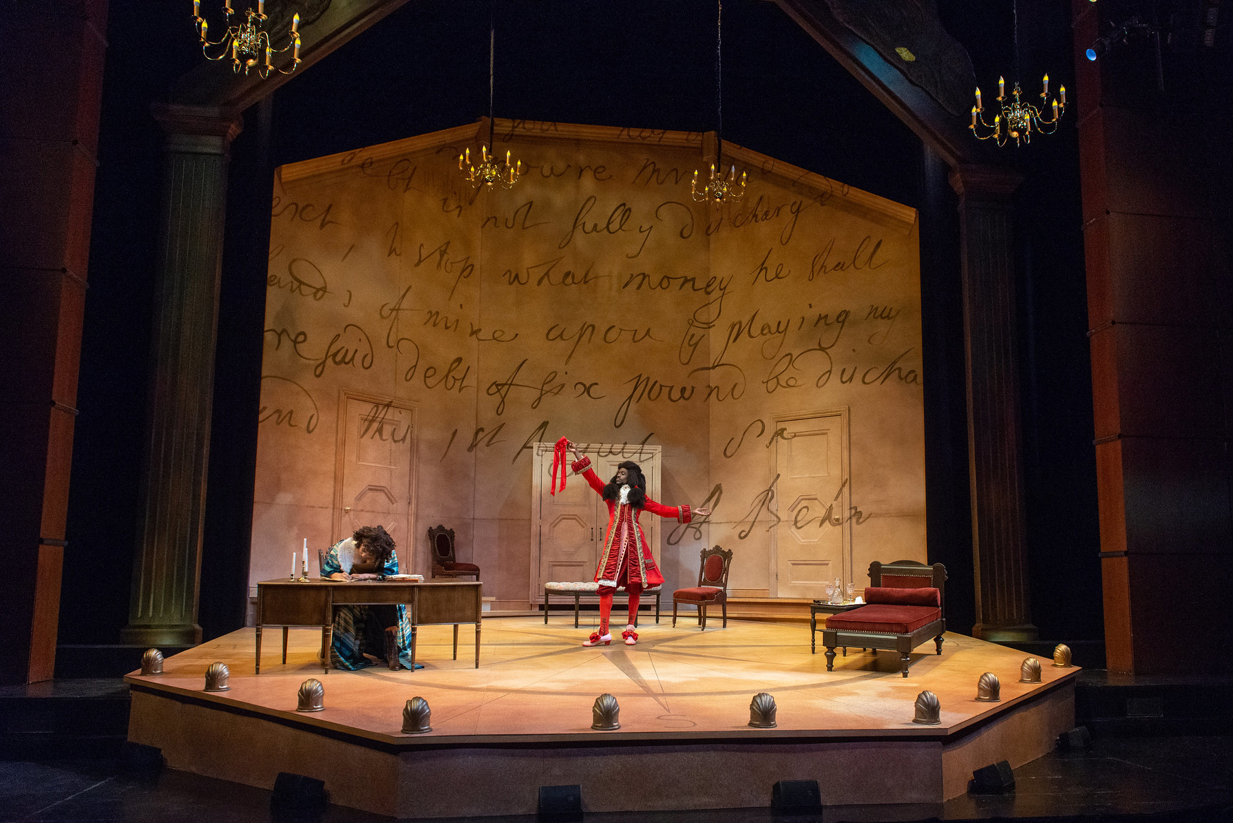 An actor center stage on a grand set, wearing a decadent red victorian coat and wig, gestures with arms outstretched; another actor in a gown sits at a table on stage, writing a letter.