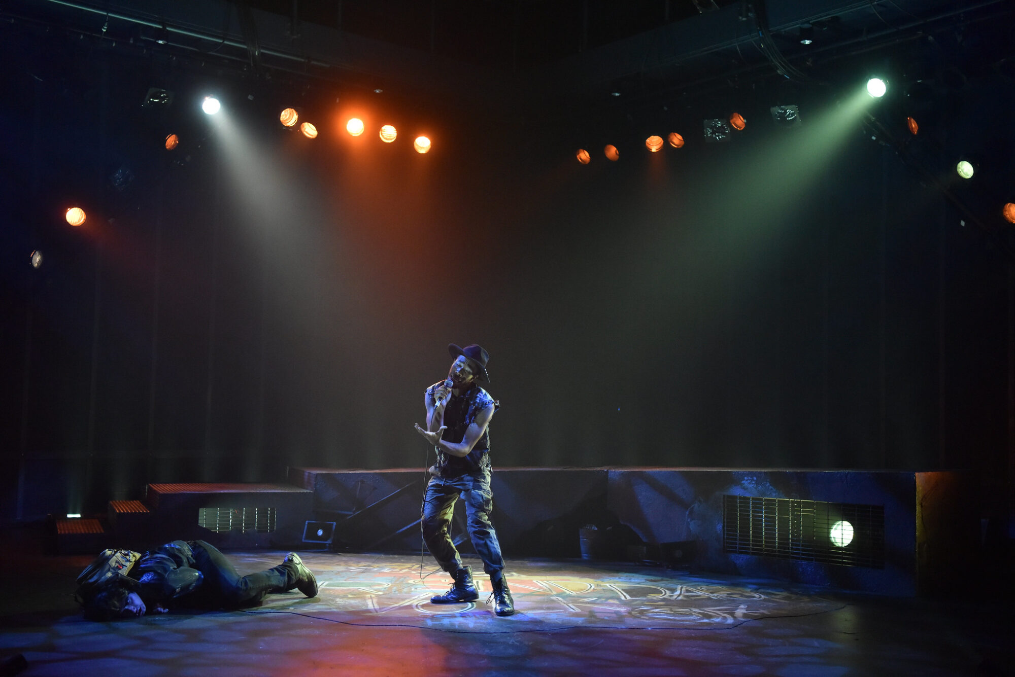 An actor sings into a standing microphone on stage, with lights shining down.