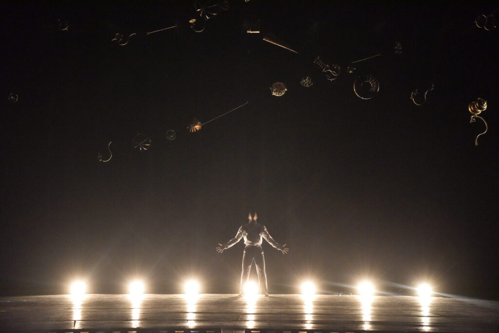 An actor stands on stage, looking up, arms outstretched; bright floor lights shine below him and various objects are suspended in the air above him.