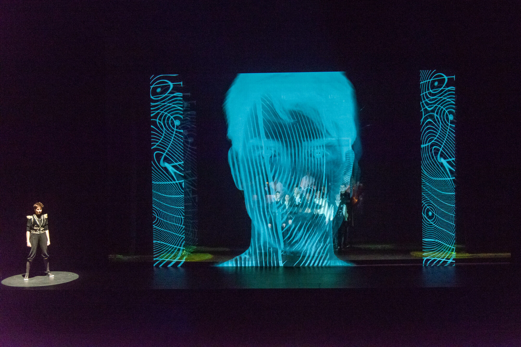 An actor stands in a spotlight on stage while a large projection of a human face fills the stage to their right.