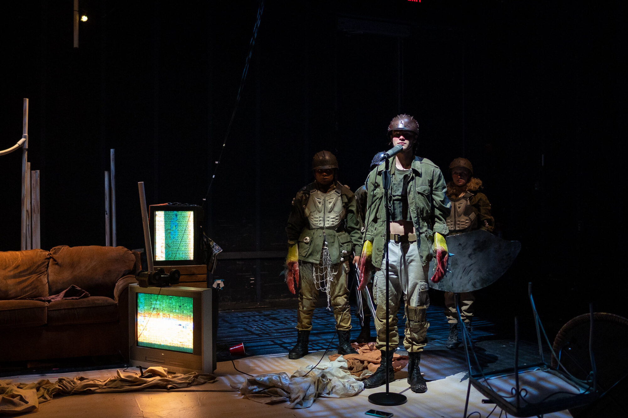 Actors dressed in army fatigues with crab claws on their hands stand at a microphone on the set of a play called “Adult Things.”