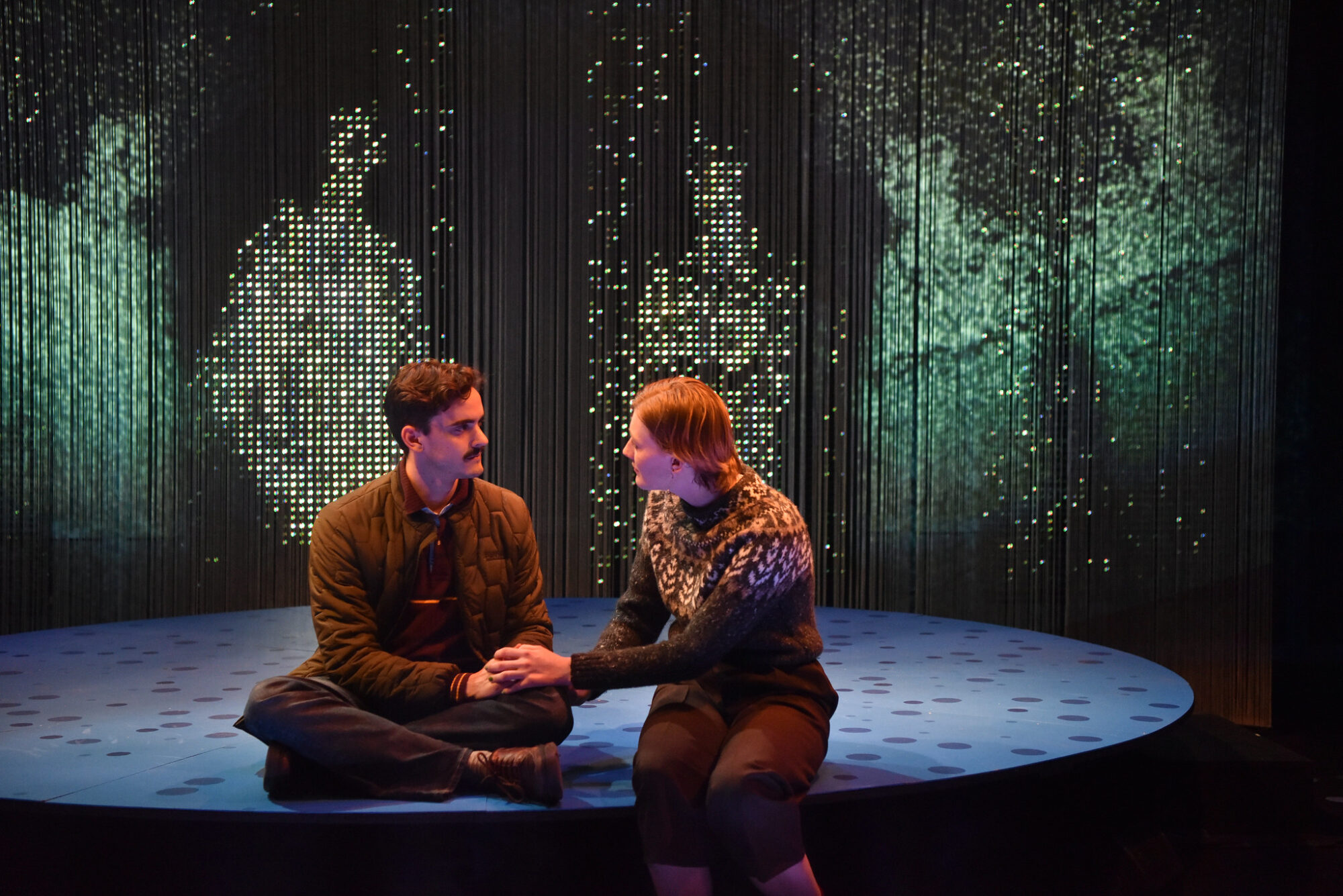 Two actors seated on stage with intricate lighting and projection depicting human forms behind them.