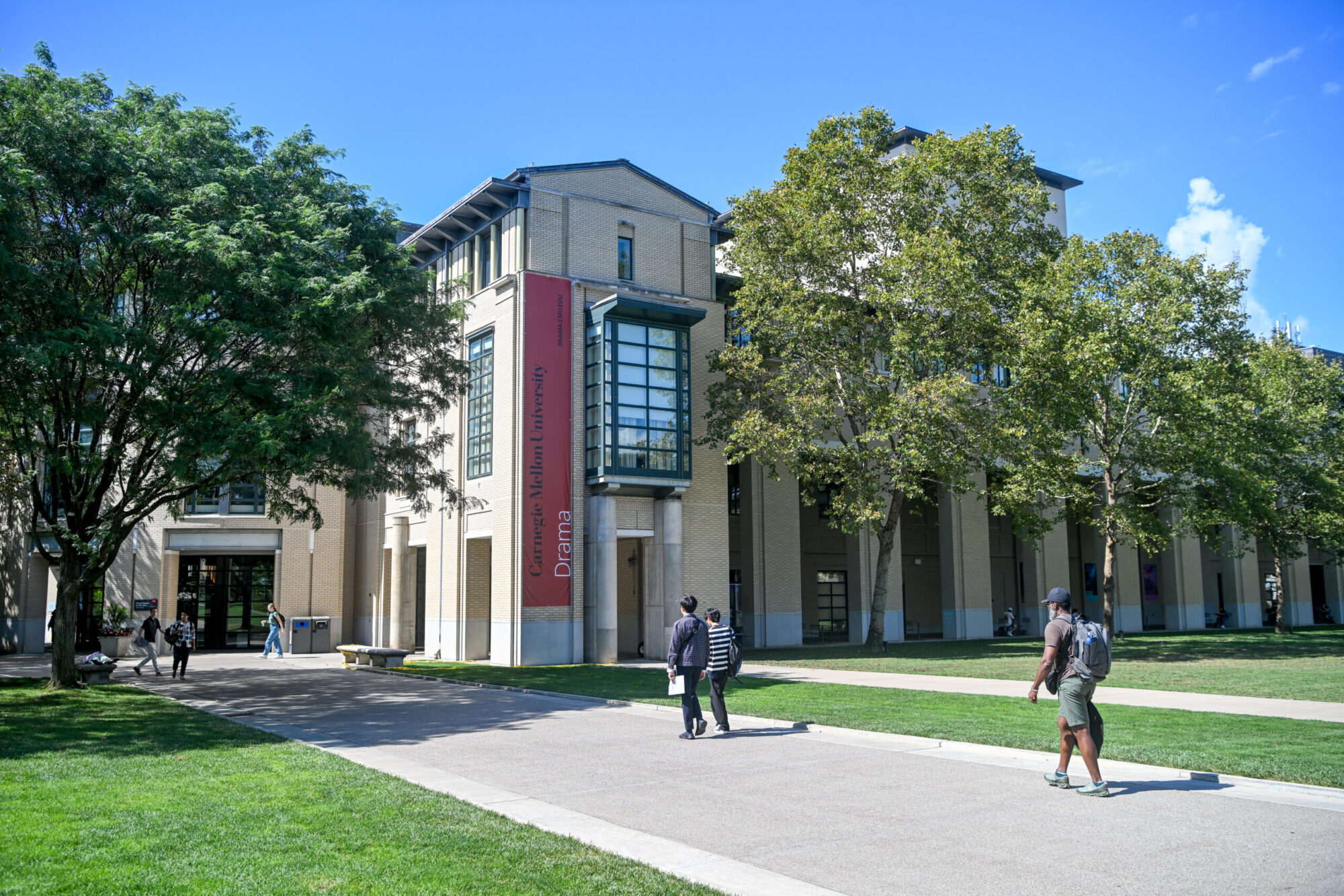 An exterior shot of the Purnell Center for the Arts, a large light colored building with a red banner on one column, surrounded by trees and green grass.