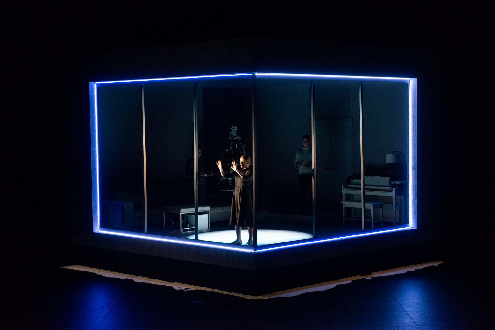 A box set of windows on all sides is illuminated on a stage with led lights around its perimeter; we see an actor inside the box in a spotlight, holding her hands up to the glass.