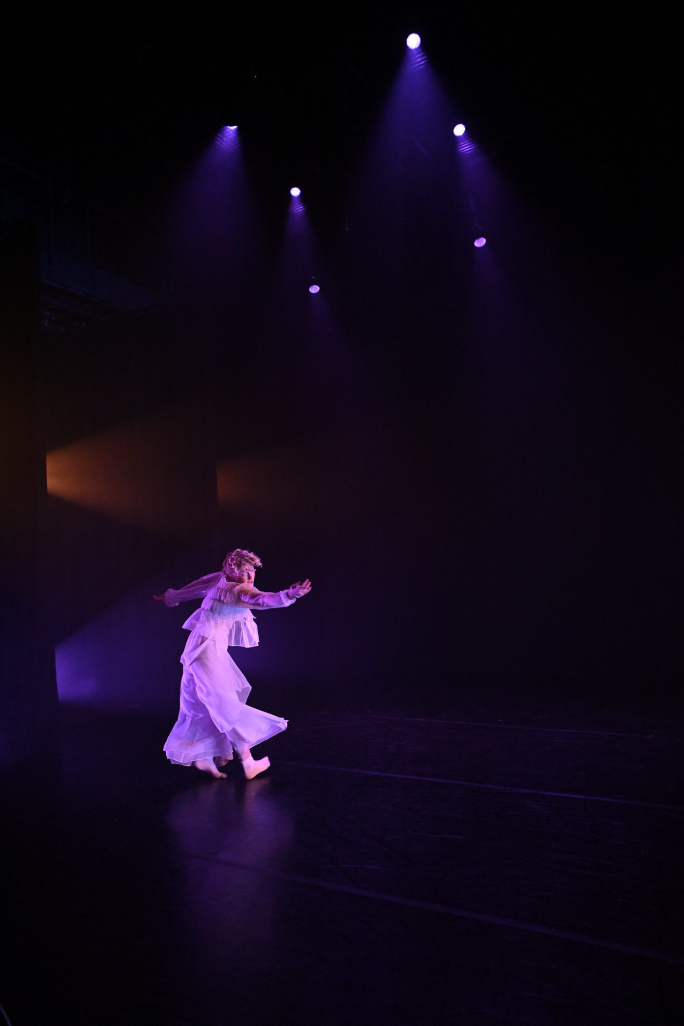 A dancer wearing all white is mid-dance, with lights illuminating overhead and from the side wings.