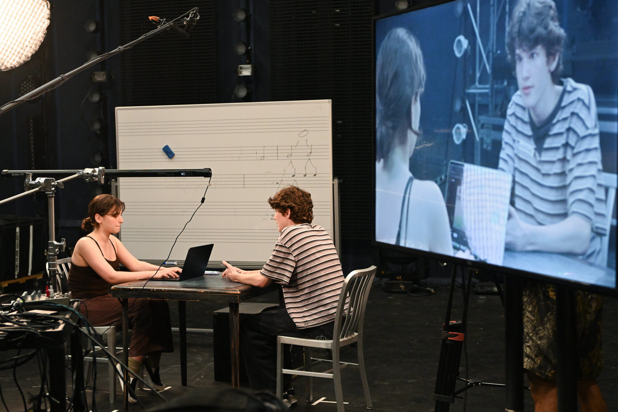 Two high school students sit at a table, acting a scene, while it is being filmed and projected onto a screen to the side of them.