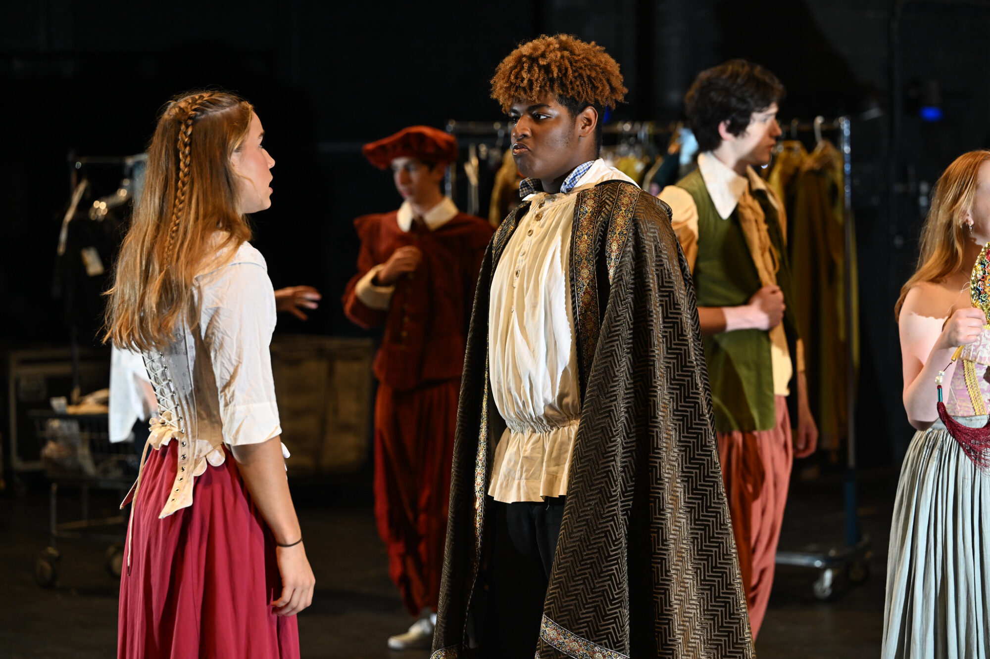 High school students face each other on stage, wearing Shakespearean costumes, rehearsing scenes.