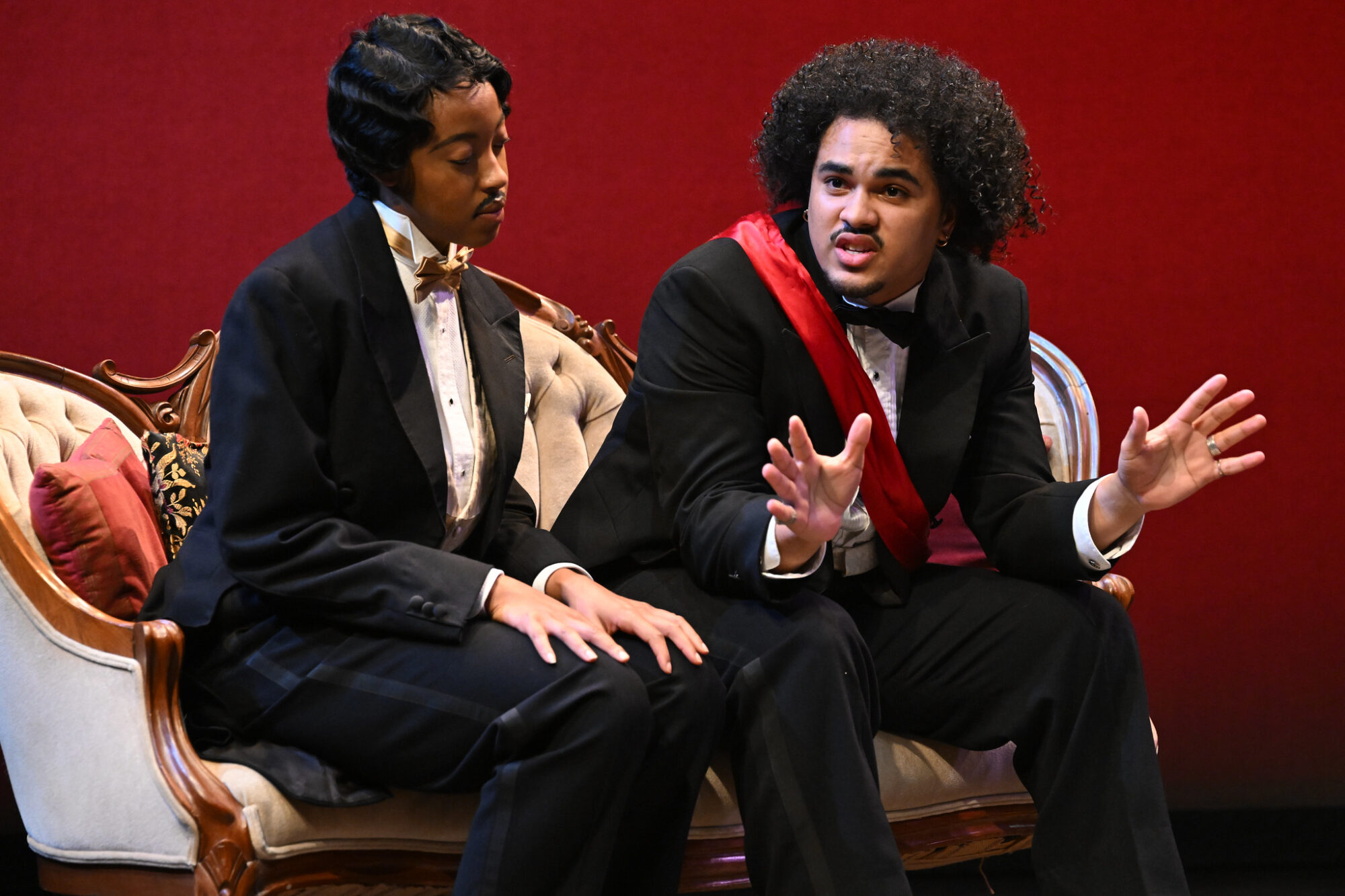 Two actors sit on a chaise on stage.