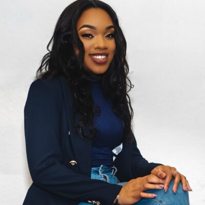 Apriah Williams, a woman with long black hair wearing a blue blazer and jeans, smiles at the camera.