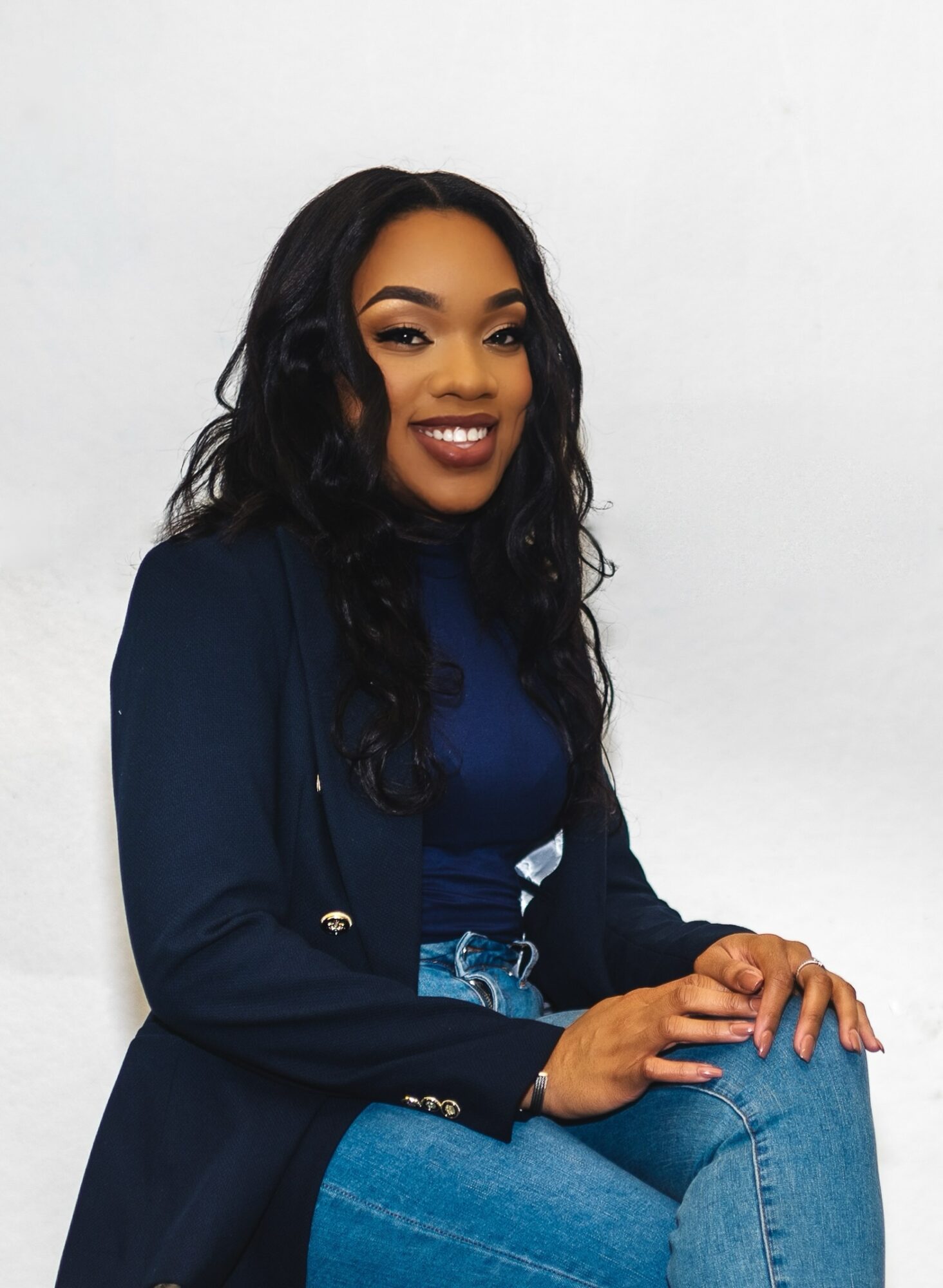 Apriah Williams, a woman with long black hair wearing a blue blazer and jeans, smiles at the camera.