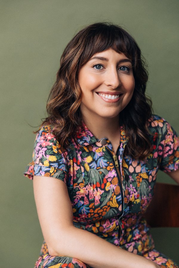 Photo of Tatiana Baccari, a woman with long brown wavy hair and bangs in a floral print jumpsuit.