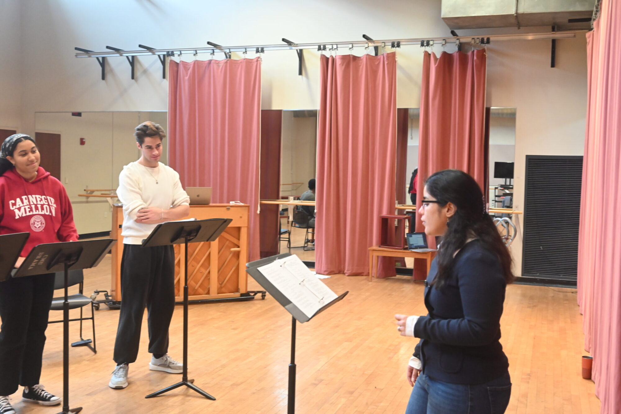 Three students standing at music stands in a rehearsal studio with red curtains and a piano.