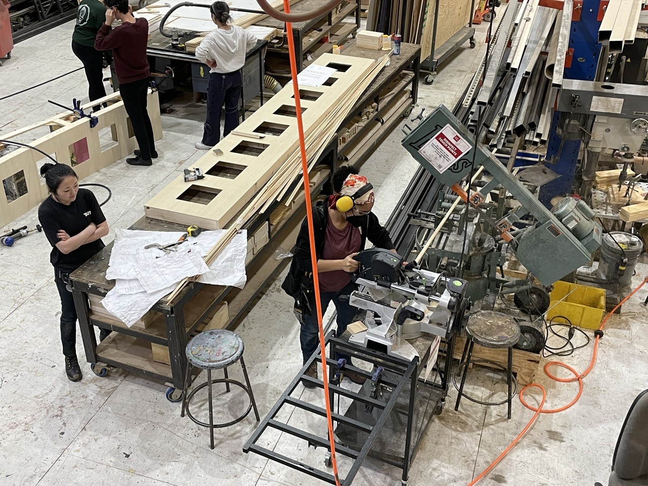 An overhead shot of the School of Drama’s scene shop, where a student wearing protective headwear, works with a table saw.