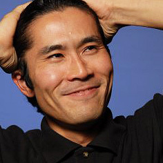 Hidenori Navajo wears a black shirt and has his hands behind his head, looking off to the side and smiling.