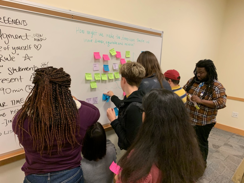 A group of students is sticking post-its to a white board, the text reads: How might we make the American theater more diverse, equitable, and inclusive?