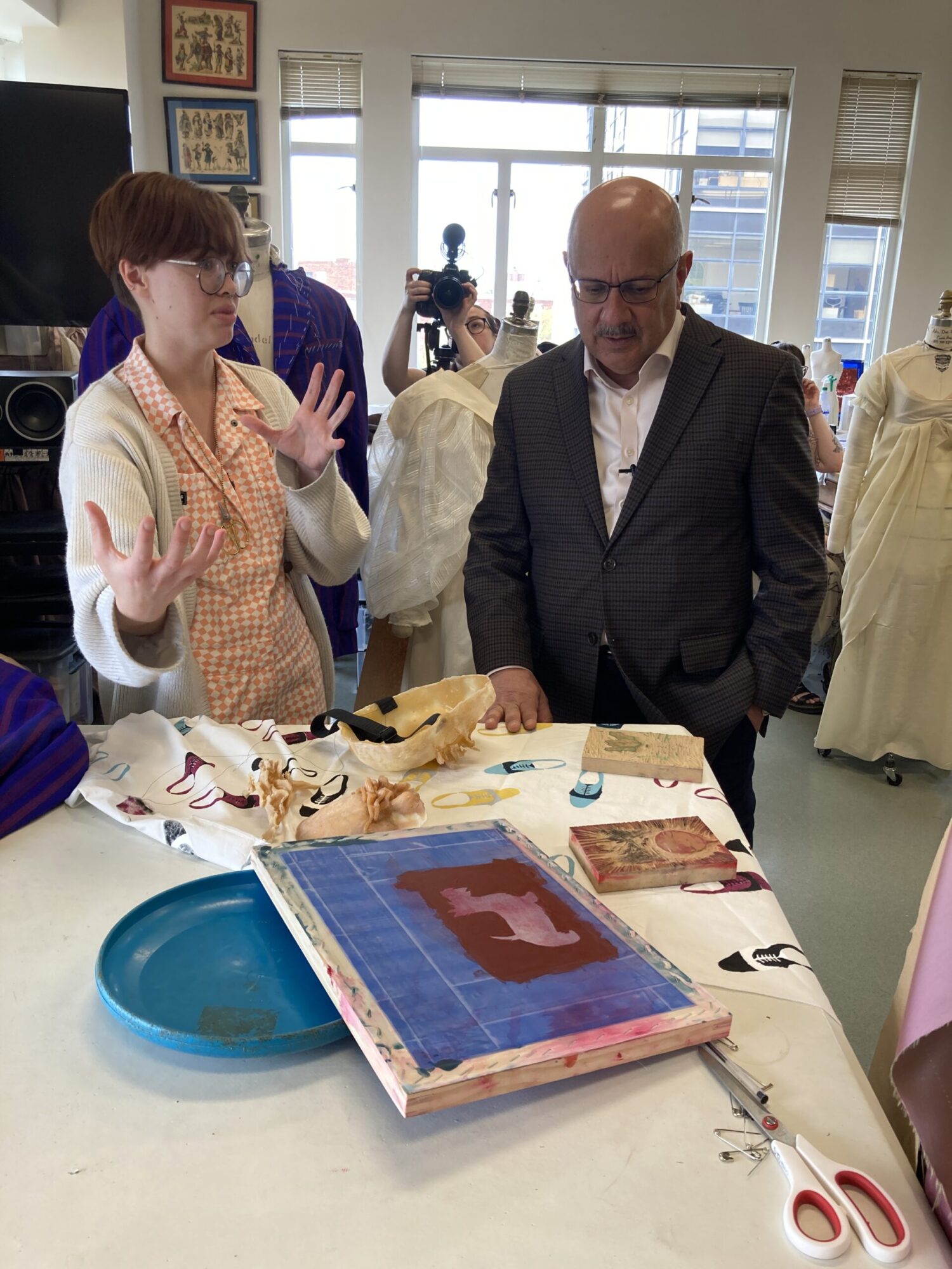 A student stands in front of a table covered in screen printing materials, gesturing in conversation with Farnam Jahanian, President of CMU, who wears a suit.