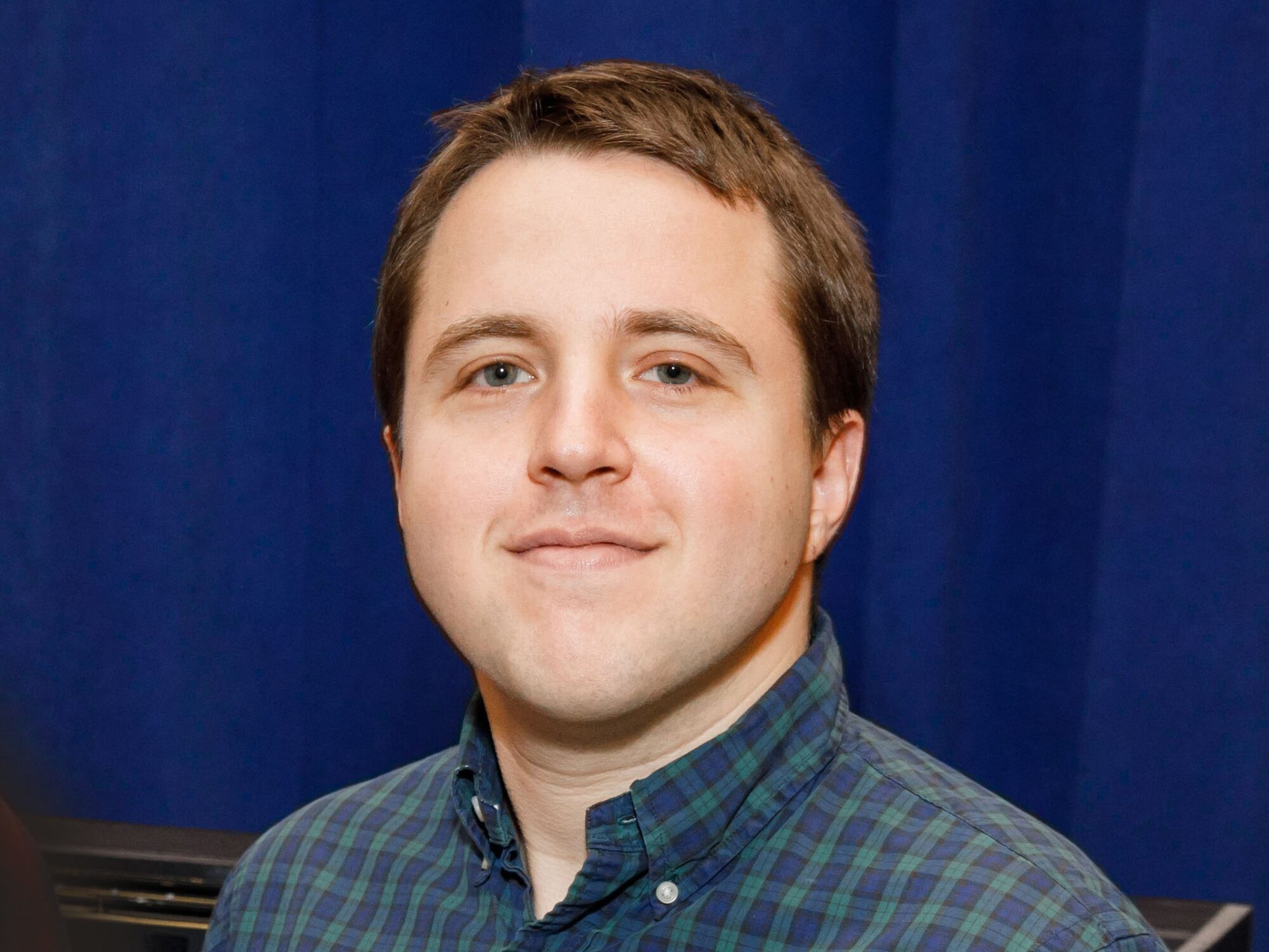 Joshua Harmon smiling at the camera, wearing a blue and green Tartan patterned collared shirt.