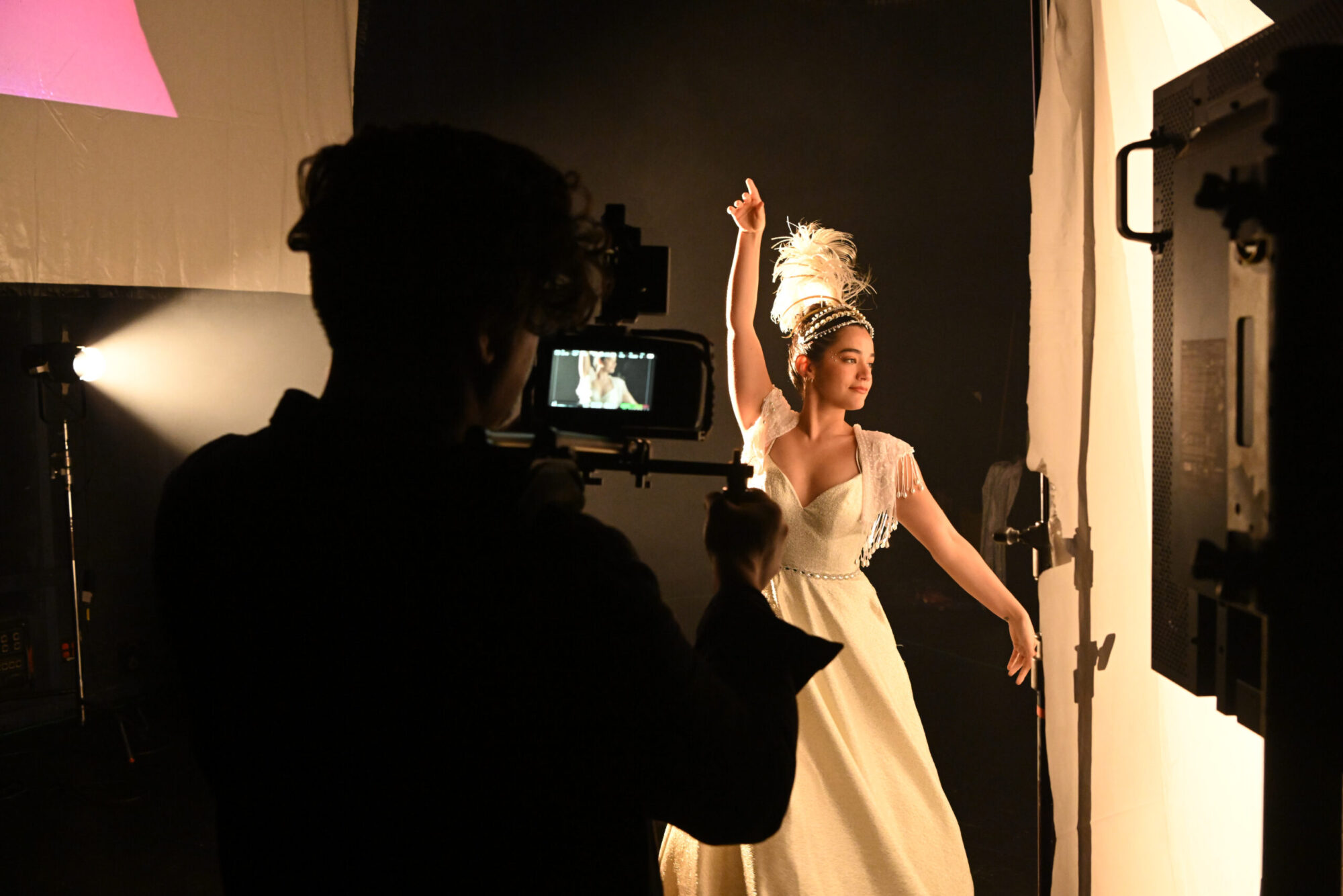 An actor in a white gown and feather headband strikes a pose behind a white screen; a man with a camera films her from the side.