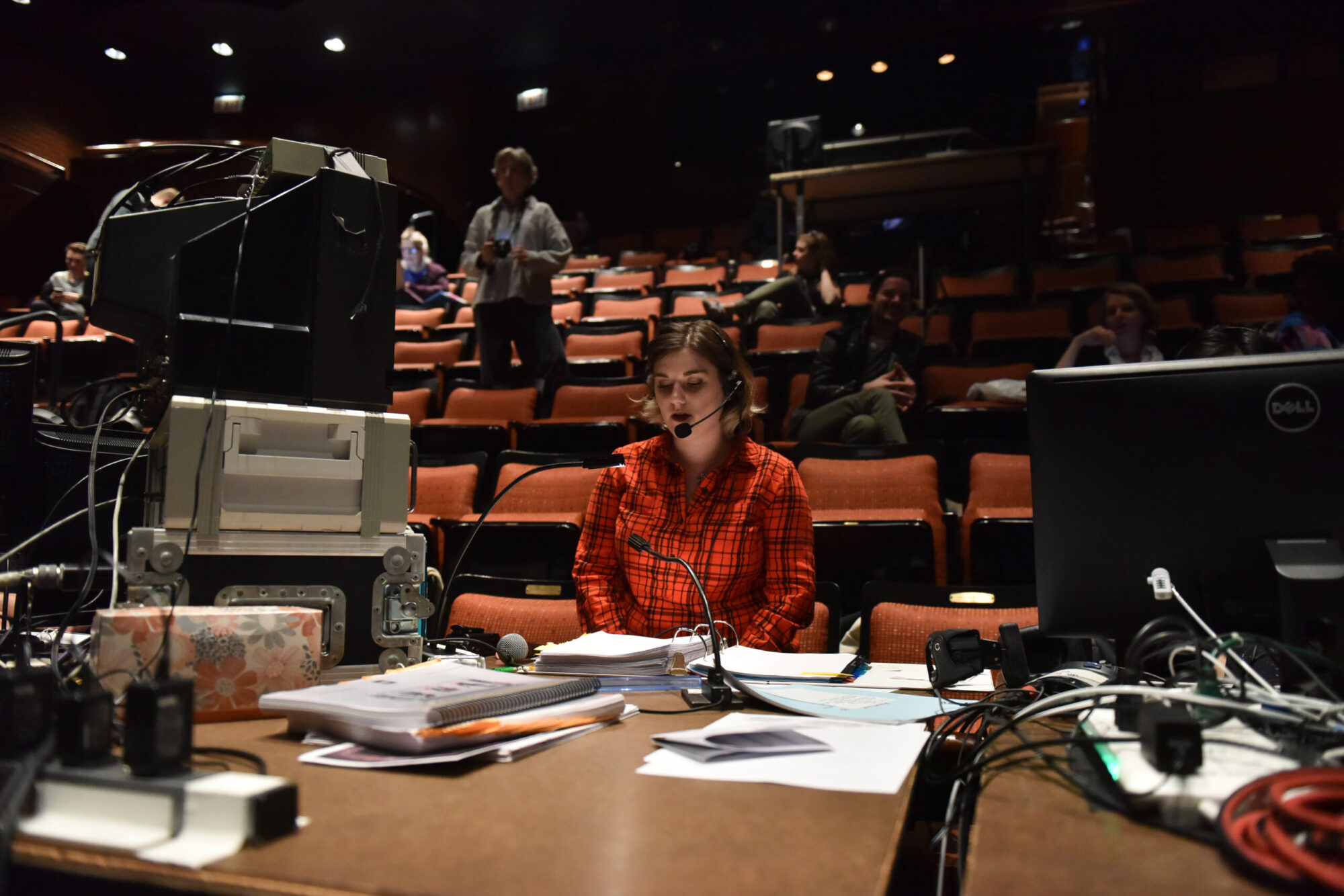 A student wearing a red shirt and a headset sits at a table inside a theater with computer monitors and a large binder open in front of her.