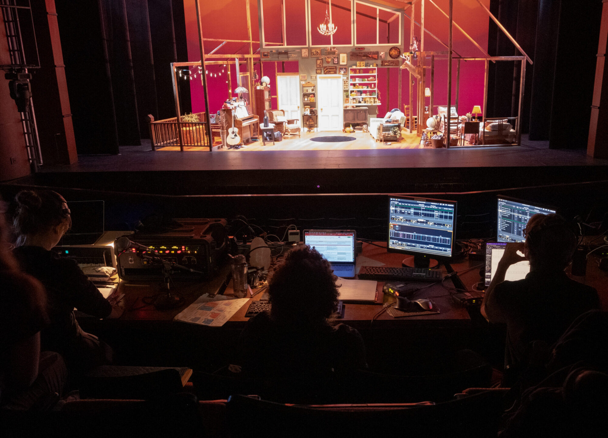 In the foreground, students are sitting at a table with computer screens lit up; in the background, we see a stage and set lit with stage lights.