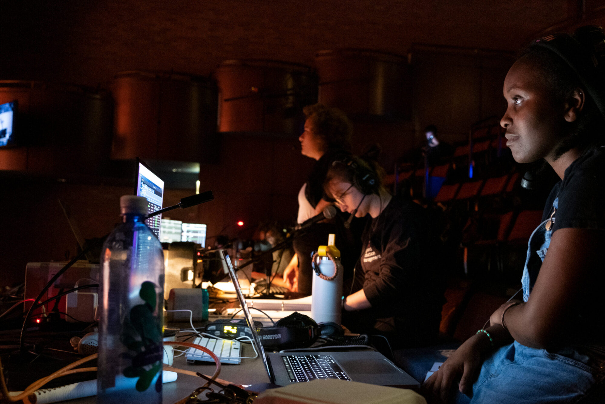 Students sit at a table in a darkened theater with computer screens and light and sound boards, working during a technical rehearsal at CMU.