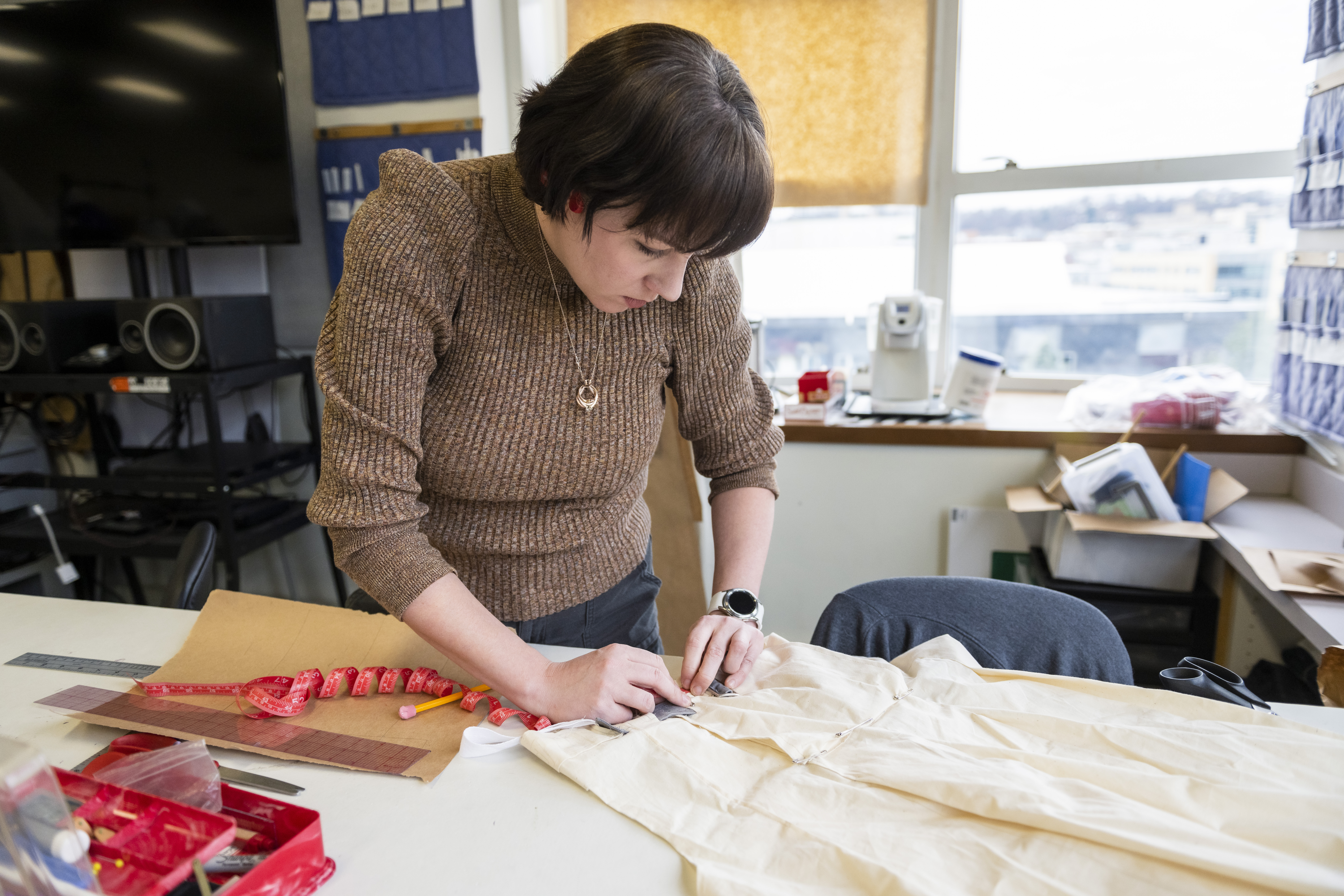 A student with short, dark brown hair, wearing a brown sweater stands at a table in the costume studio, working with her hands on a piece of muslin fabric.