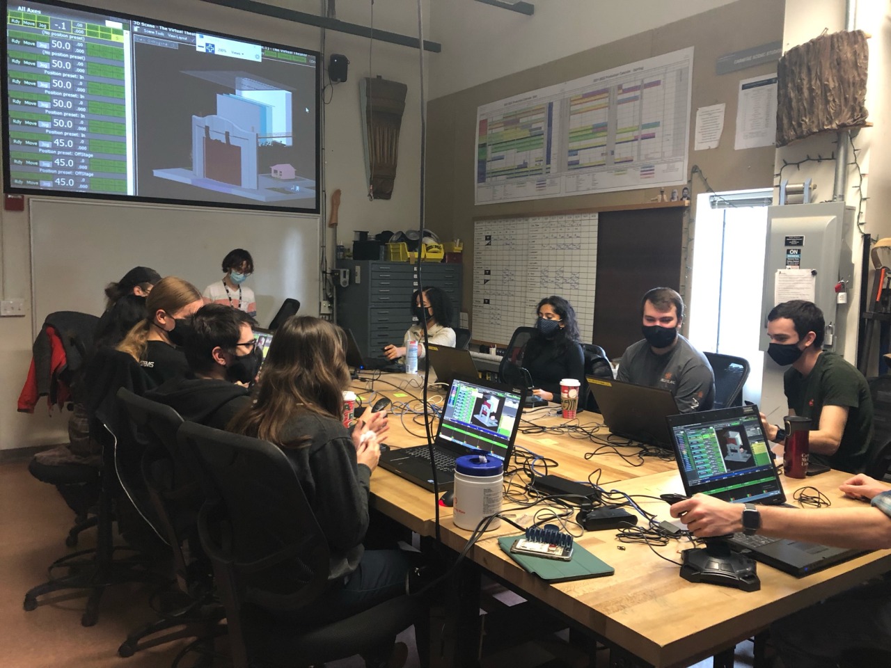 Students sit around a large desk in a classroom with laptops; a large screen on the wall shows technical software.