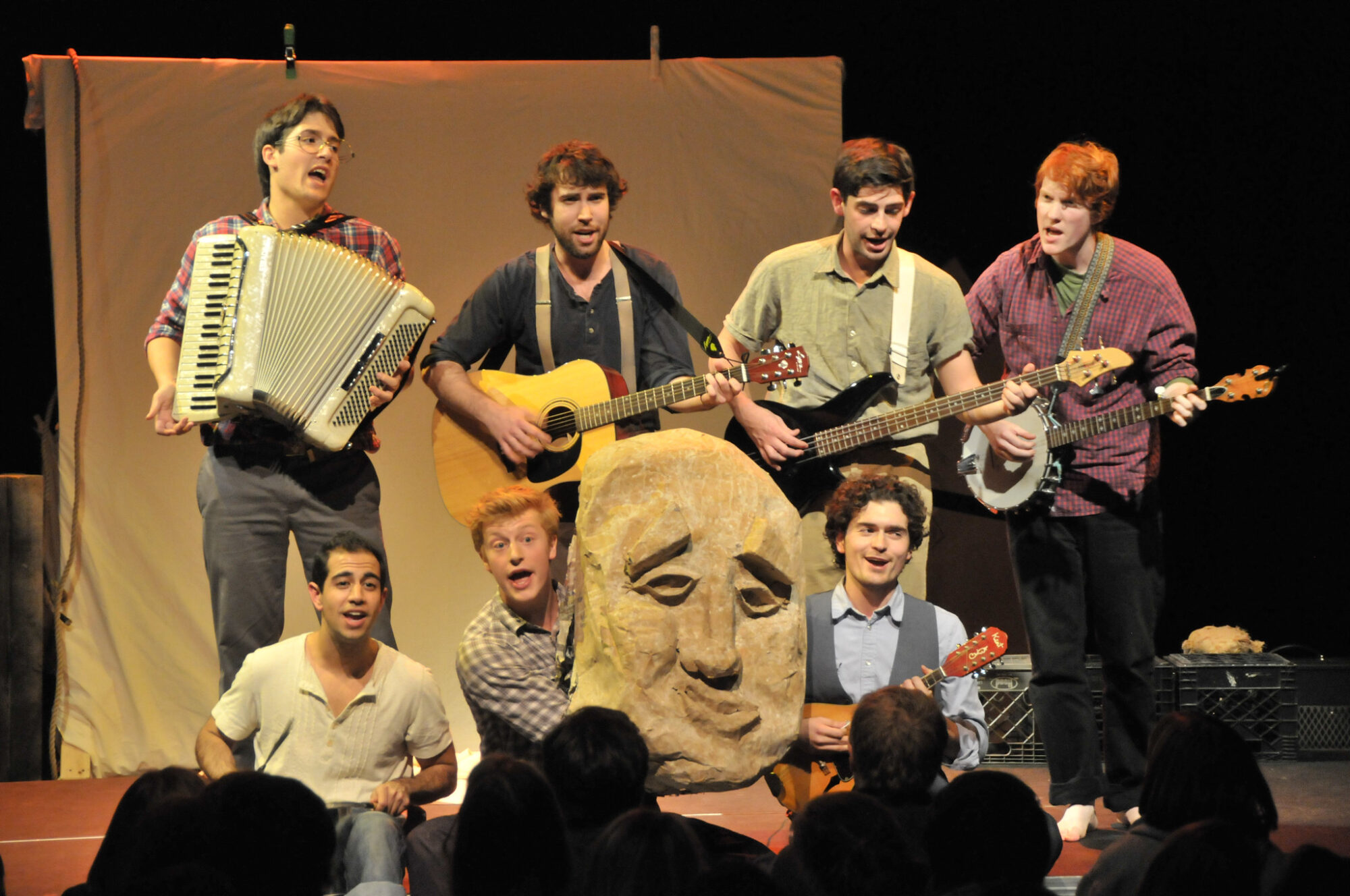 Photo of the seven members of PigPen Theatre Co. playing instruments, with a large mask in the center of the group.