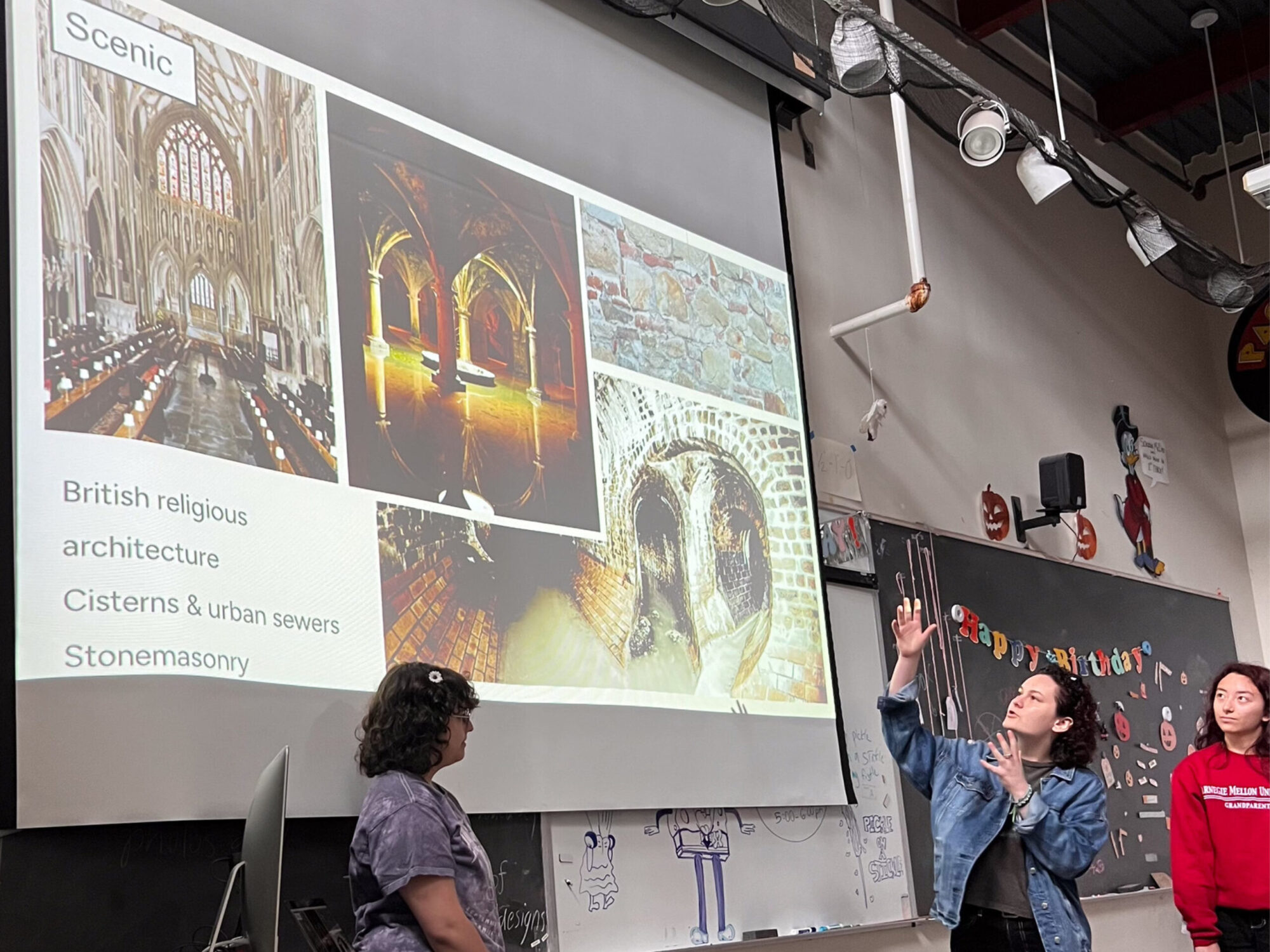 Three high school students giving a presentation, gesturing to a screen that says “Scenic” with several architectural photos.