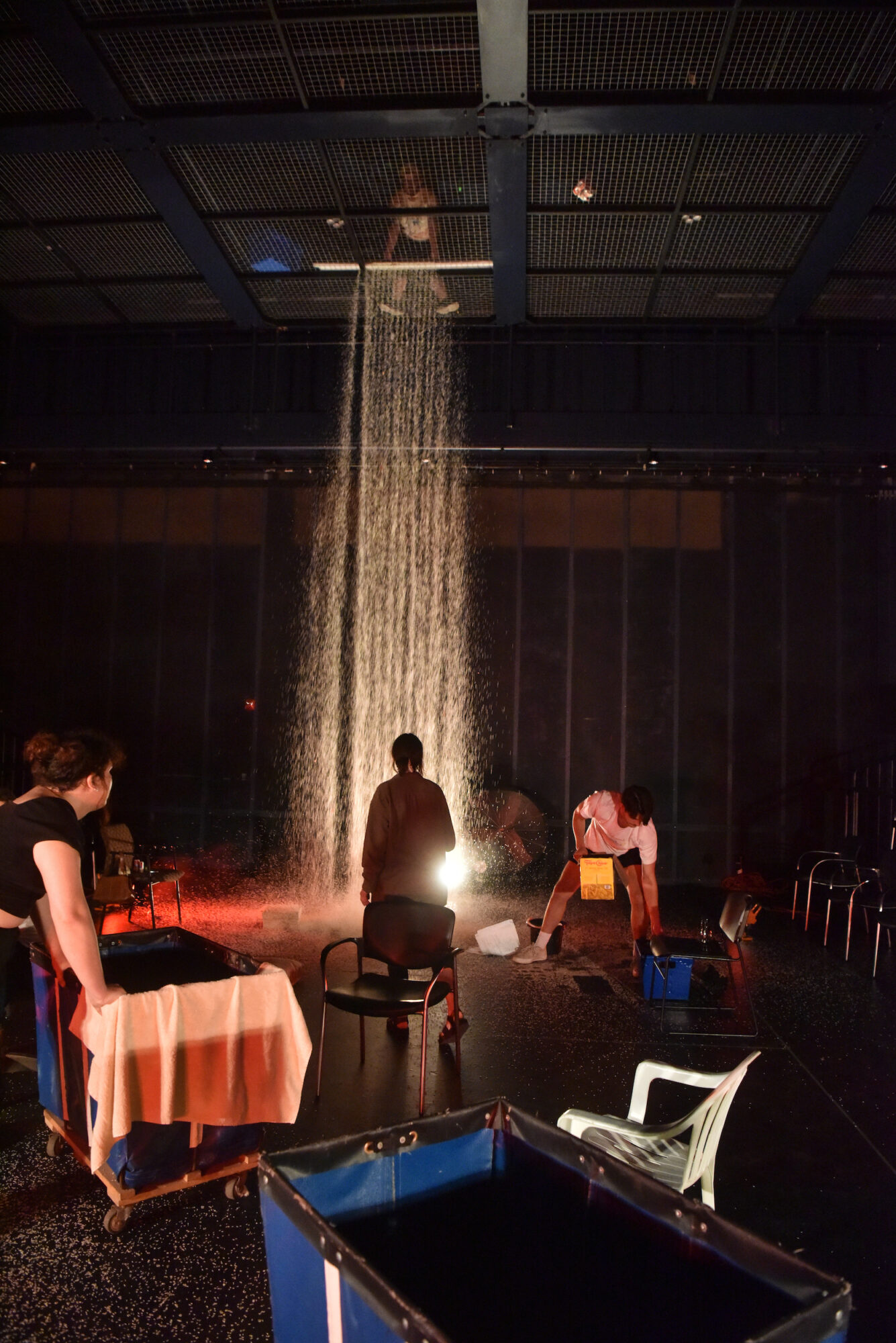 In a black box theater, rice falls from the ceiling with actors on stage