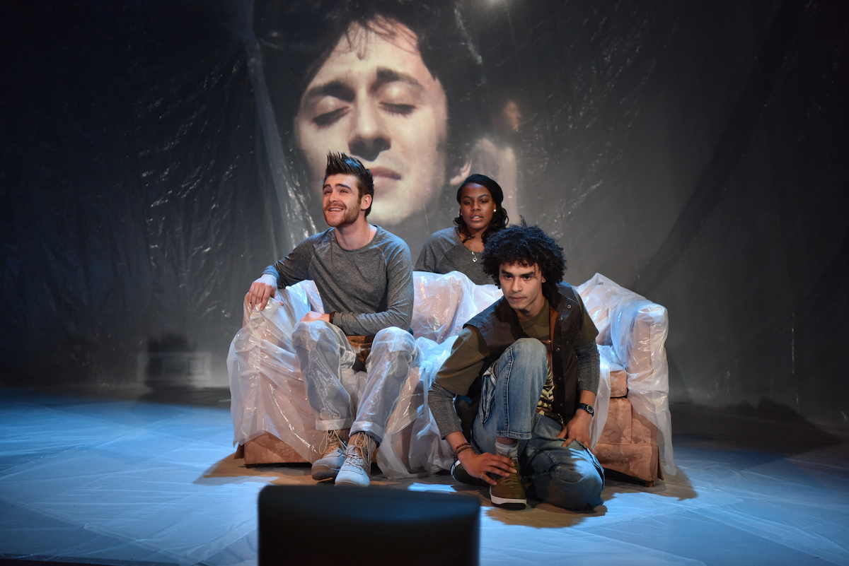 Three actors sit on furniture on stage, watching a TV screen on the ground, a face is projected on the wall behind them.