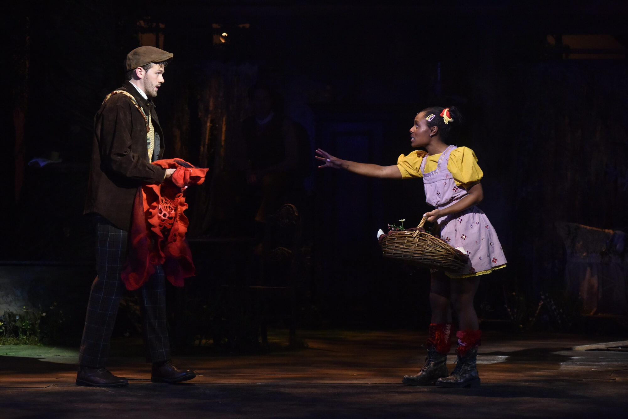 A man on stage holds a red cape; a woman on stage holds a basket, reaching for the cape, in a scene from "Into the Woods."