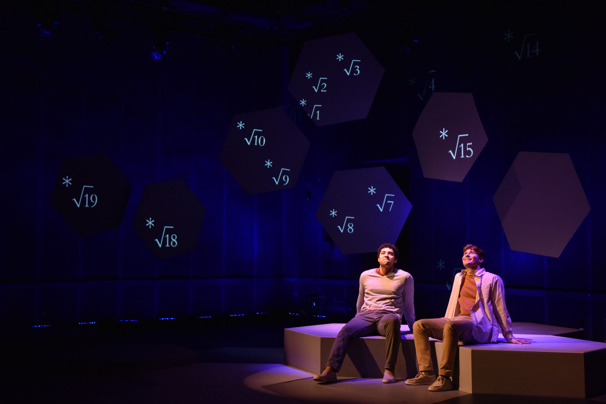 Two actors sit on stage in light, looking up, projections of mathematical symbols are projected on objects behind them.