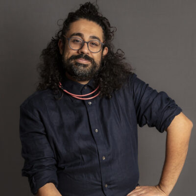 Photo of Adil Mansoor, a man with long dark hair pulled half up and a dark beard, wearing a navy blue button up shirt, seated with one hand on his hip.