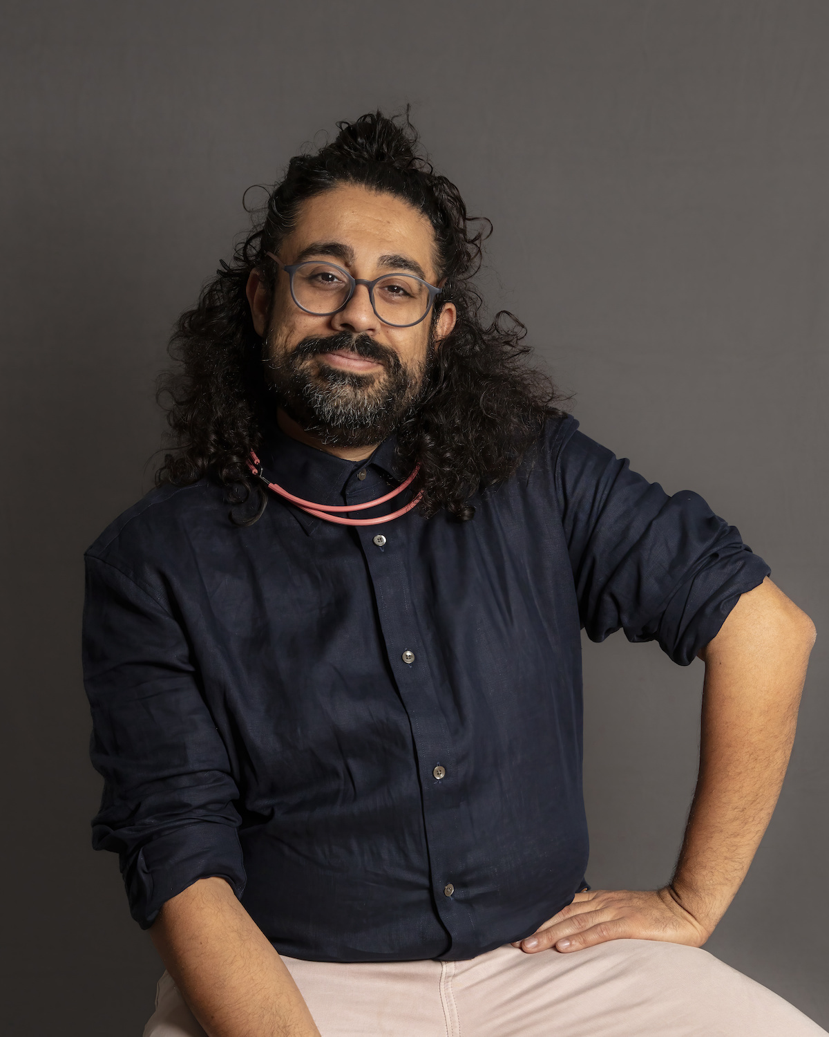 Photo of Adil Mansoor, a man with long dark hair pulled half up and a dark beard, wearing a navy blue button up shirt, seated with one hand on his hip.