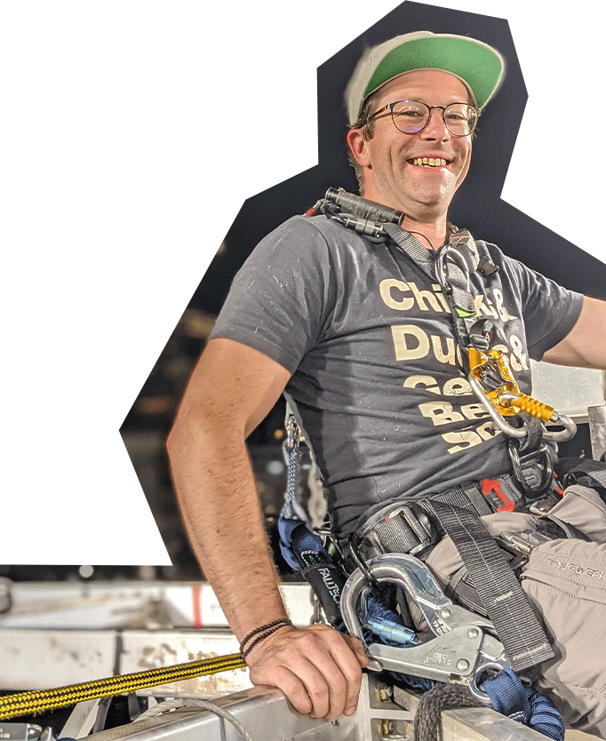 Photo of Zachary Brown, a white man with glasses, wearing a hat and t-shirt, with sound equipment, sitting atop rigging equipment and smiling.